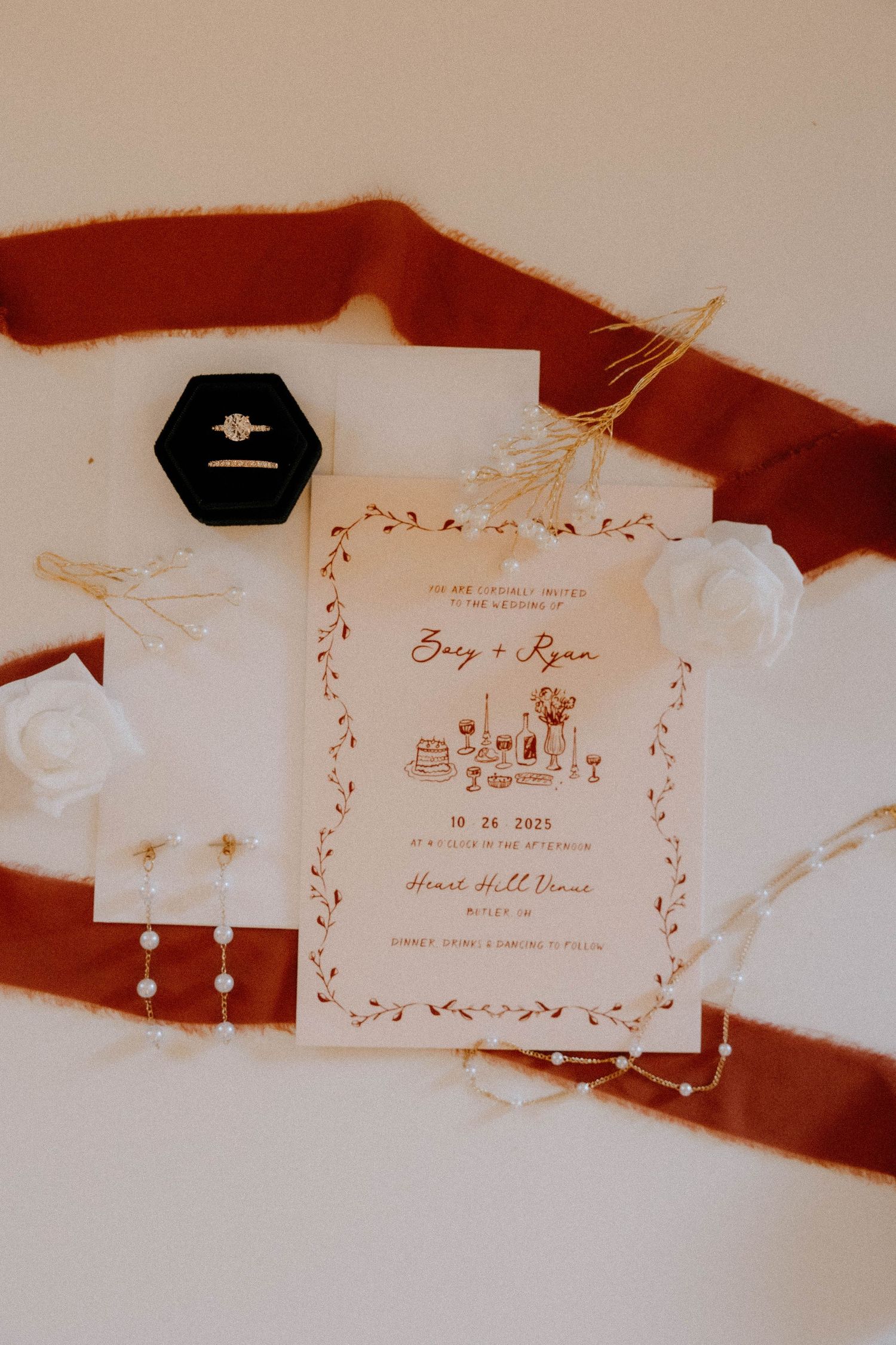 Wedding invitation with floral design and black ring box on terracotta fabric background.