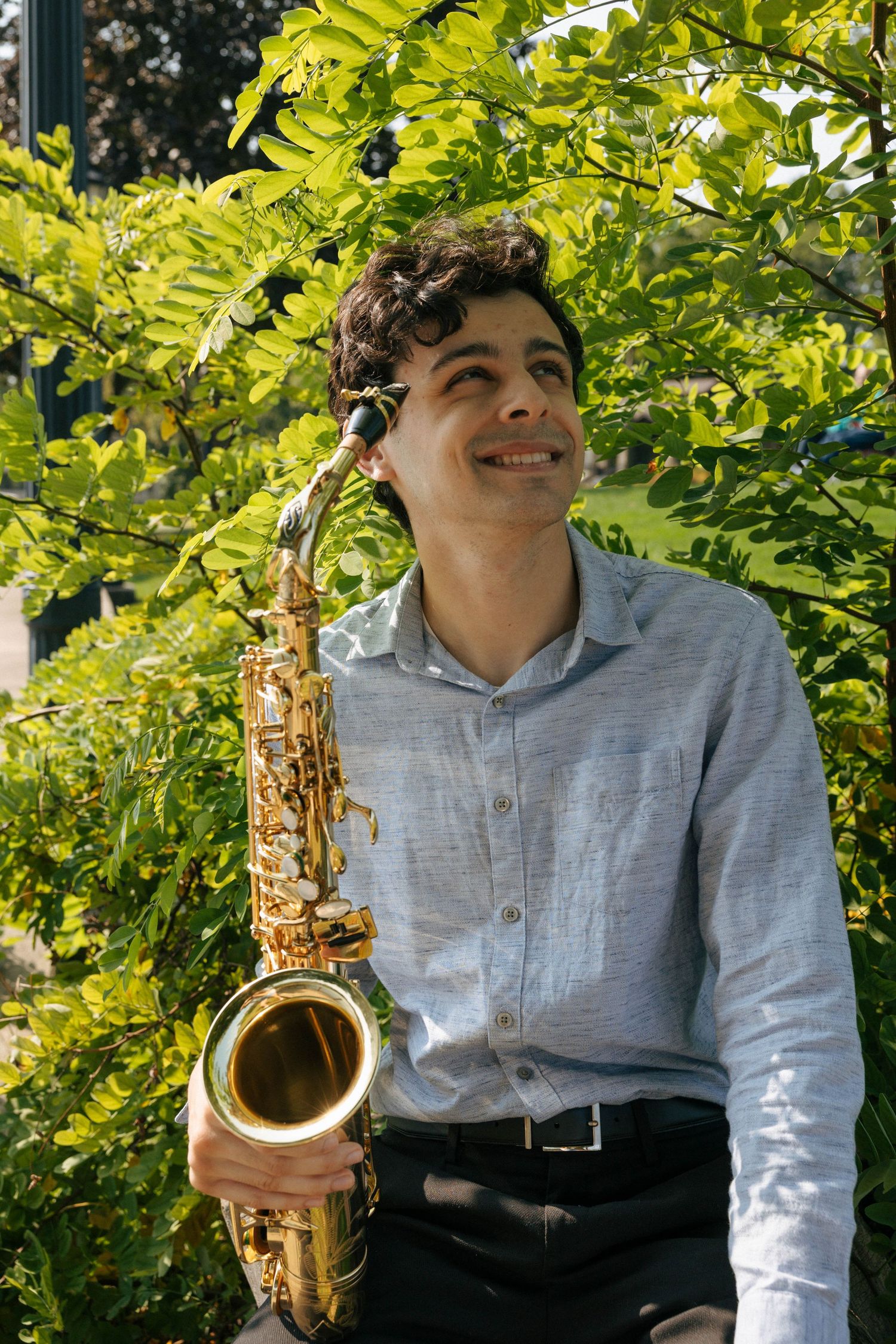 Musician with saxophone posing against lush green foliage background.