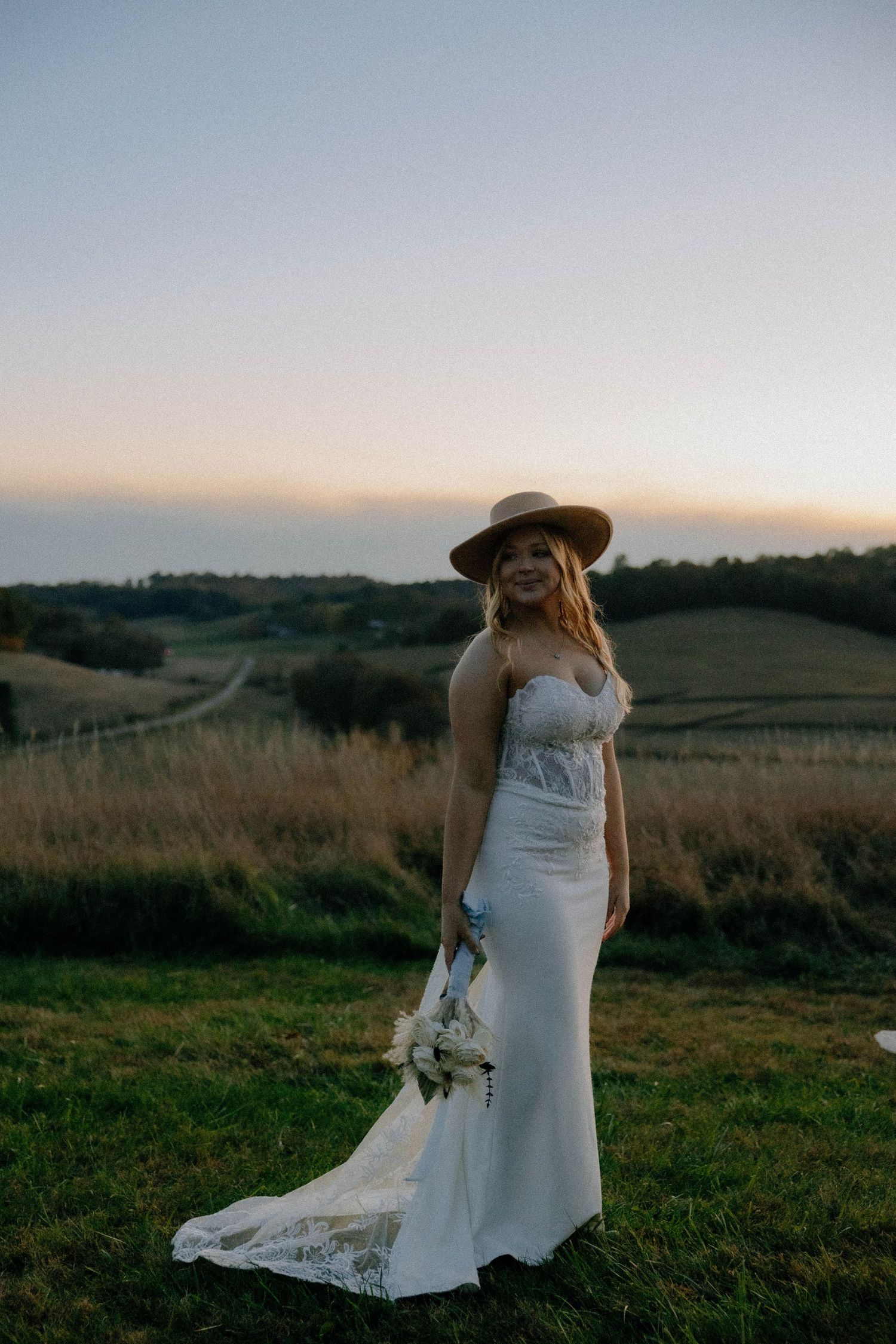 A bride in a lace wedding gown stands against a backdrop of rolling countryside hills.