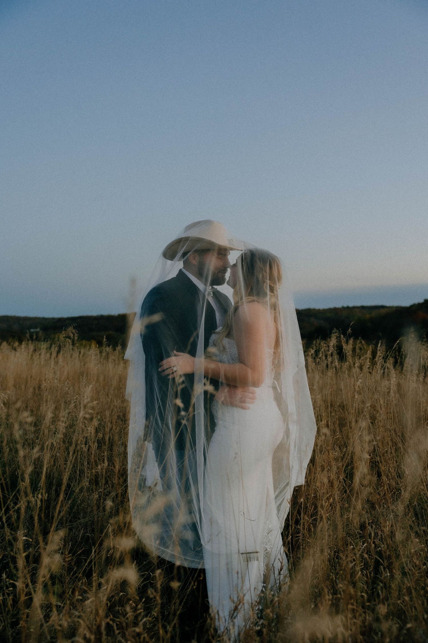 A romantic couple embraces in a golden wheat field at sunset, with the bride wearing a flowing white veil.