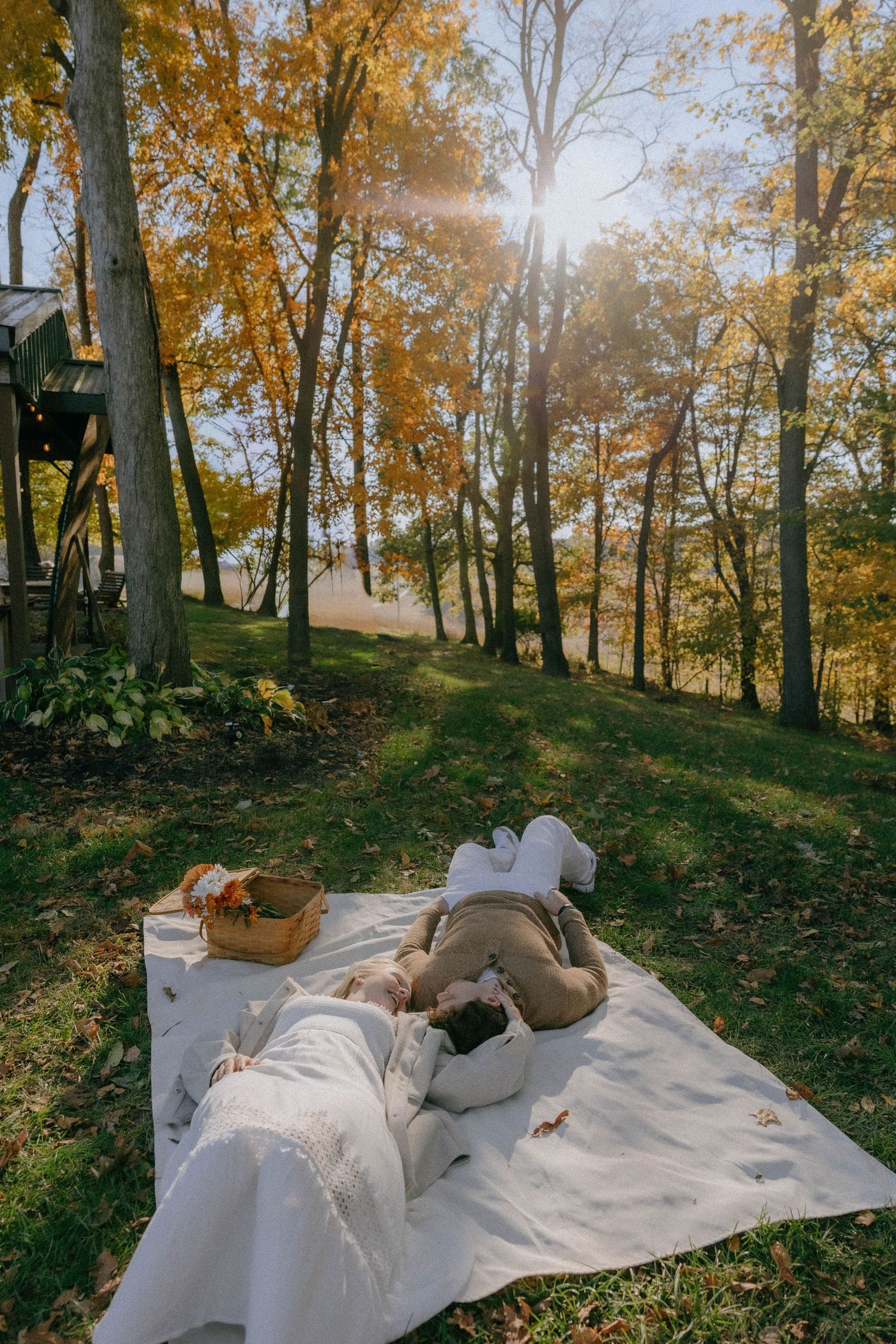 A cozy autumn picnic setup with a white blanket spread on fallen leaves in a wooded area.