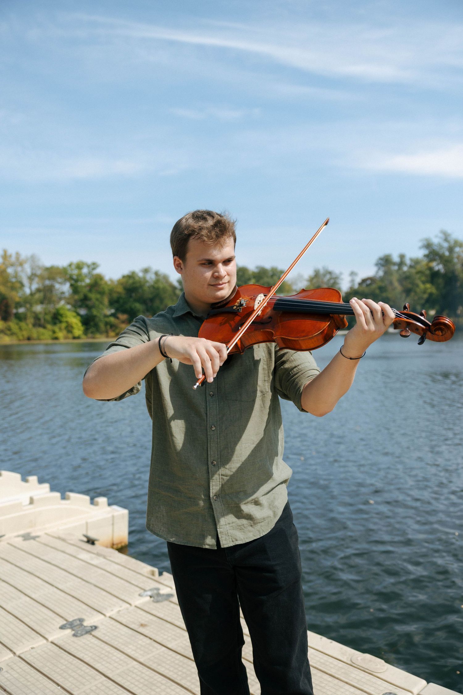Musician playing violin on a wooden dock by a serene lake on a sunny day.