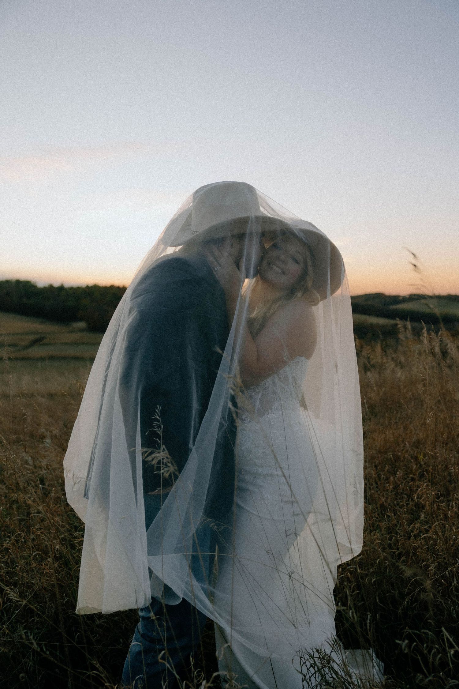 Artistic silhouette of newlyweds embracing under a flowing veil at sunset in a field.