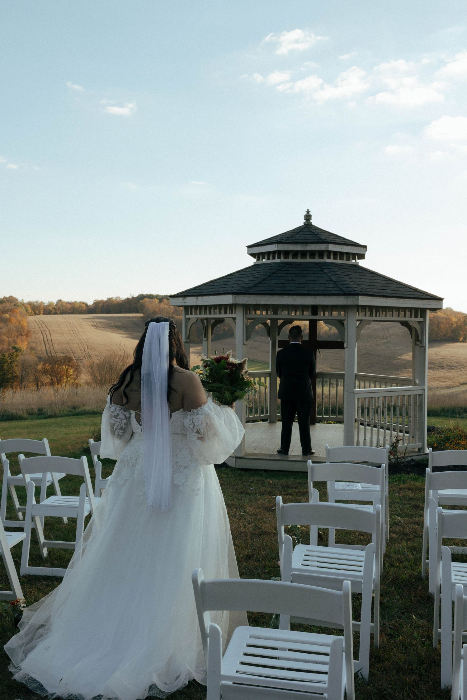 Outdoor wedding ceremony setup with white chairs and a gazebo against a scenic natural backdrop.