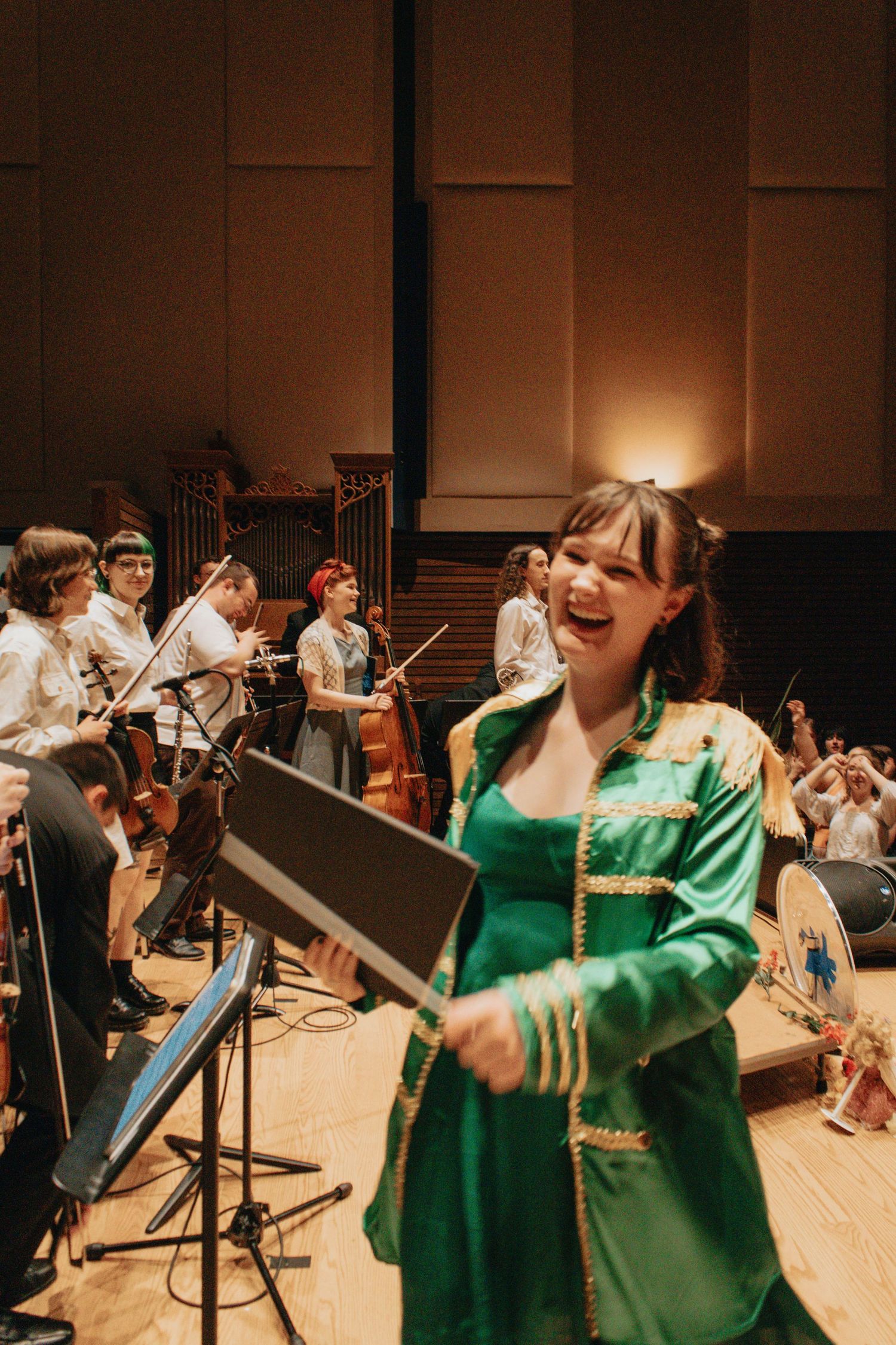 Musicians perform in formal attire on stage with sheet music stands and orchestral instruments.