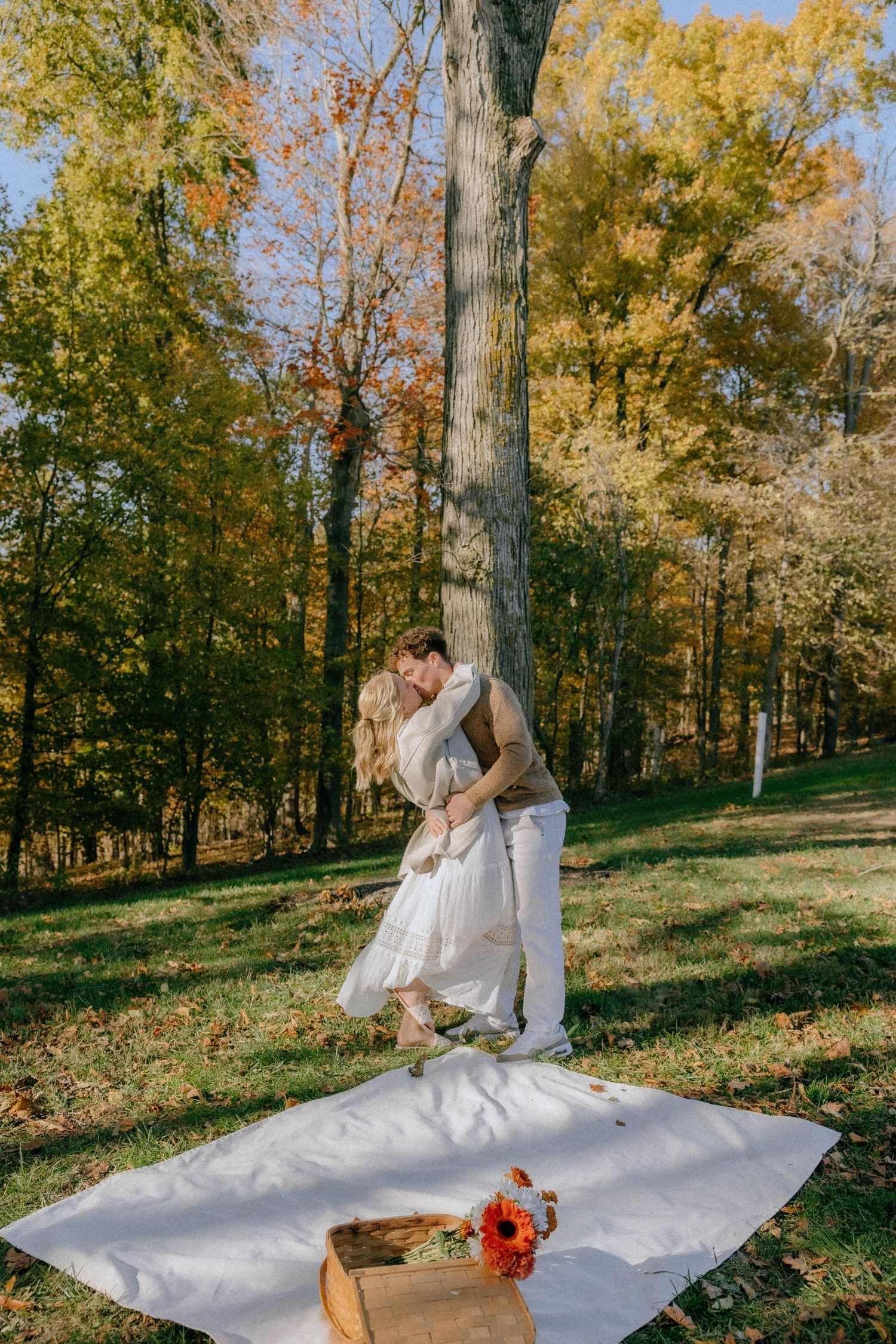 A romantic outdoor picnic setup with a white blanket and flowers under golden autumn trees.