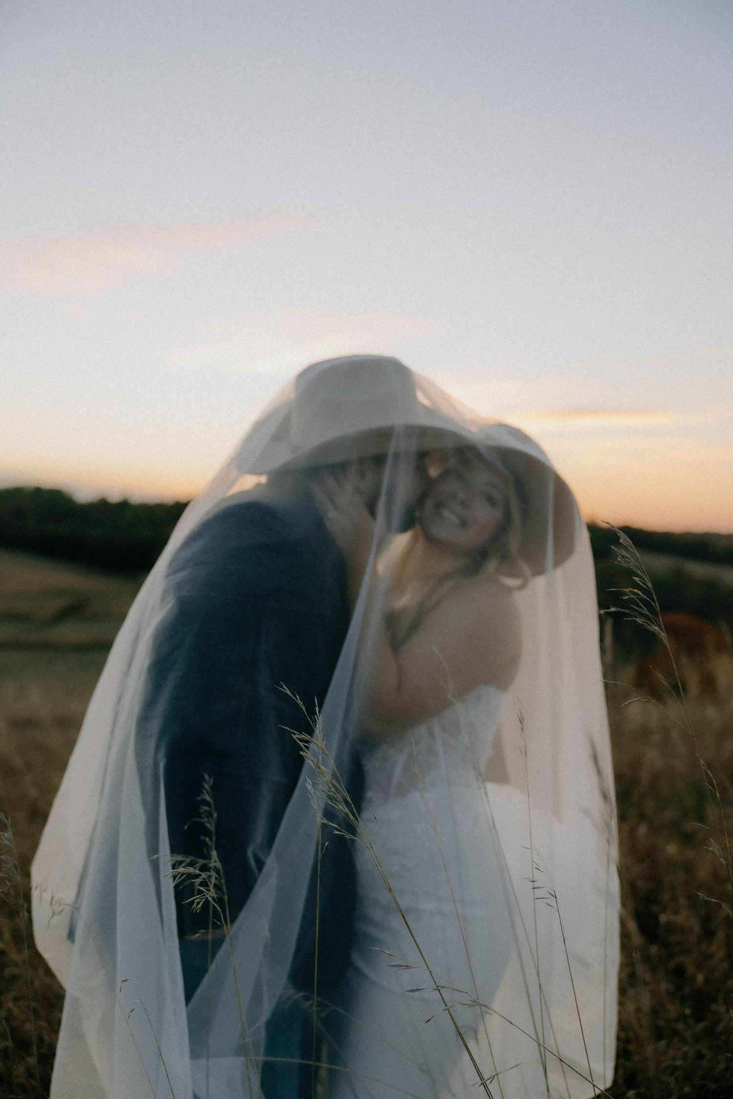 Couple embracing under a sheer veil at sunset in a golden field.