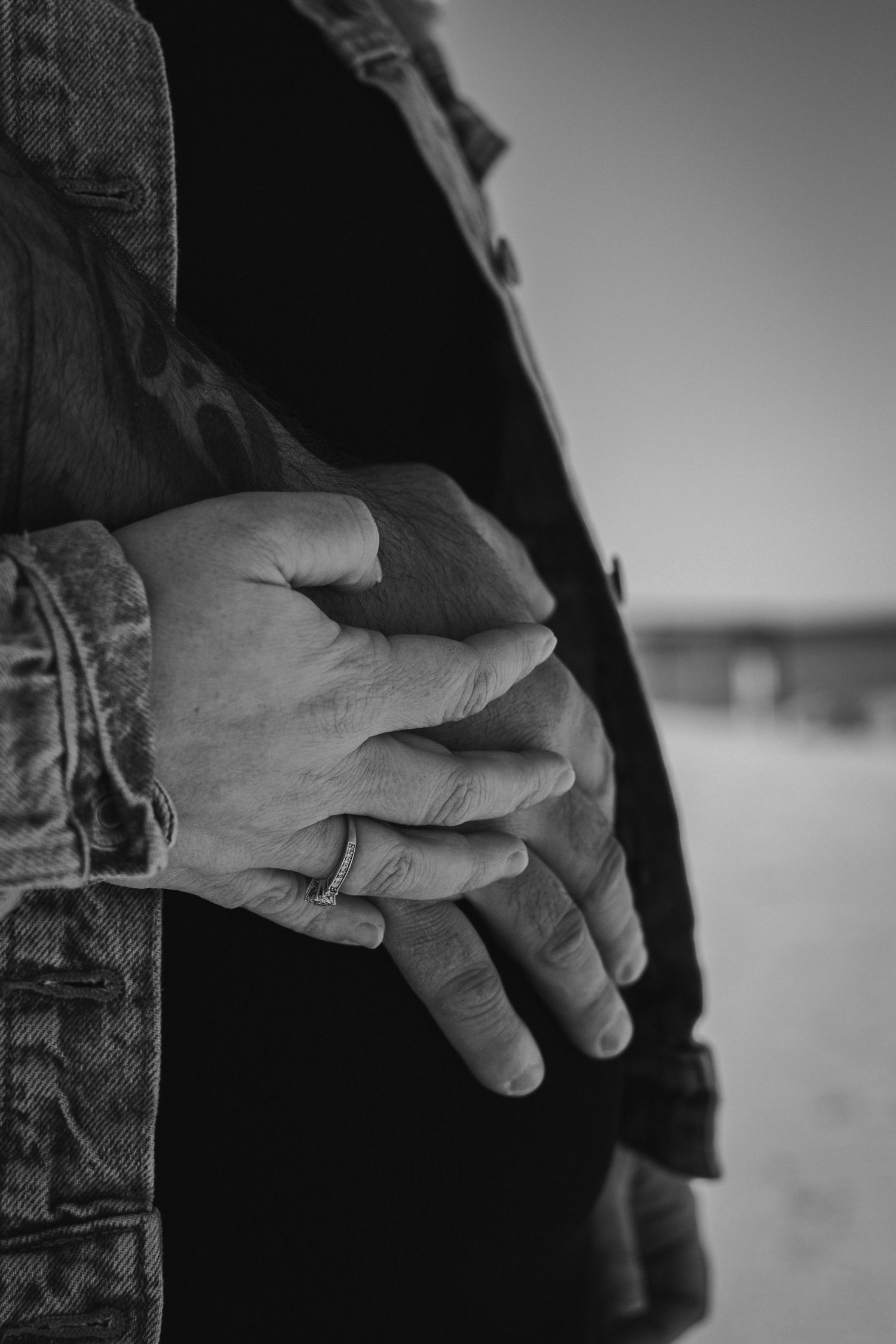 Close-up of couples hands on a pregnant belly, showing a ghost-face tattoo and ring, shot in black and white at the Irondequoit, NY pier. Mo
