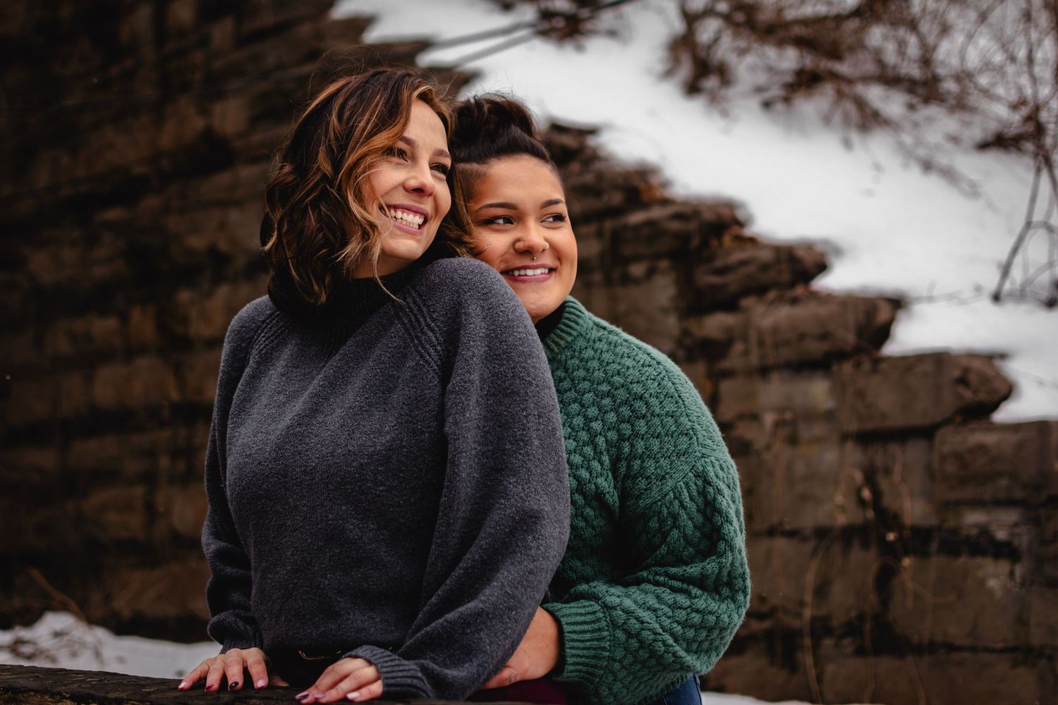 Cozy winter engagement photo of women embraced on railing at Corbett's Glen Nature Park, NY.