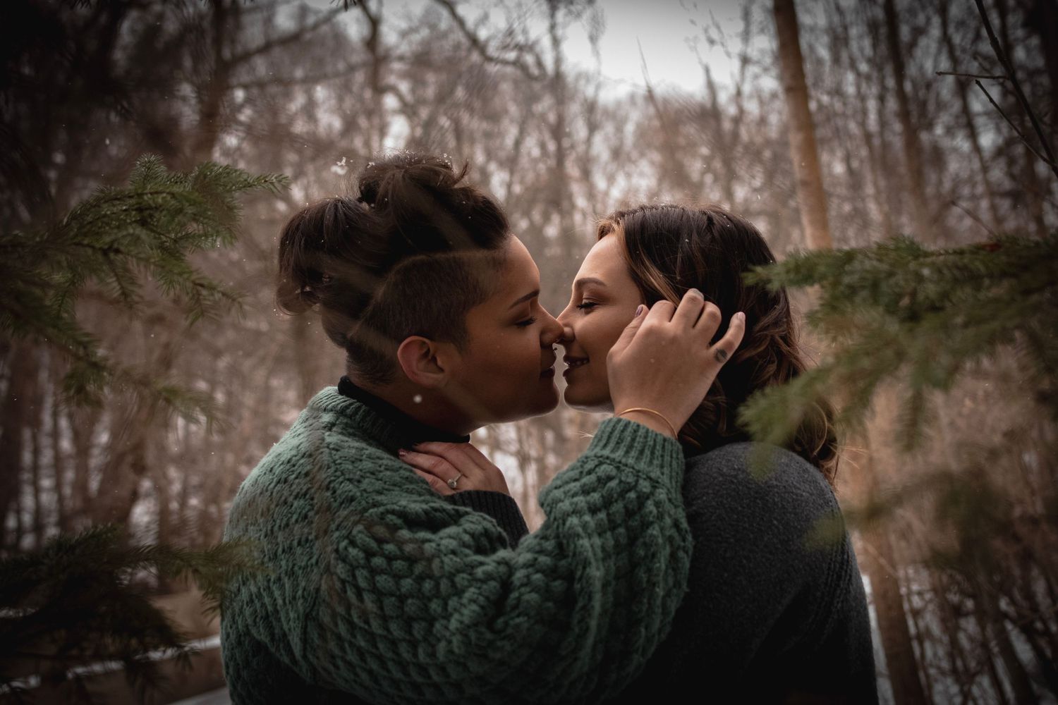 Intimate photo of two engaged women kissing under snowfall and pine trees at Corbett's Glen Nature Park, NY. Cozy winter session portrait.