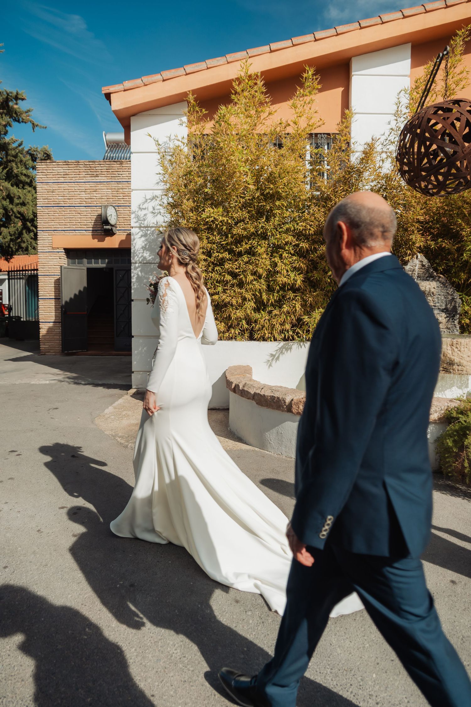 Bride in a long-sleeved white gown walks with a man in a navy suit outside a building.