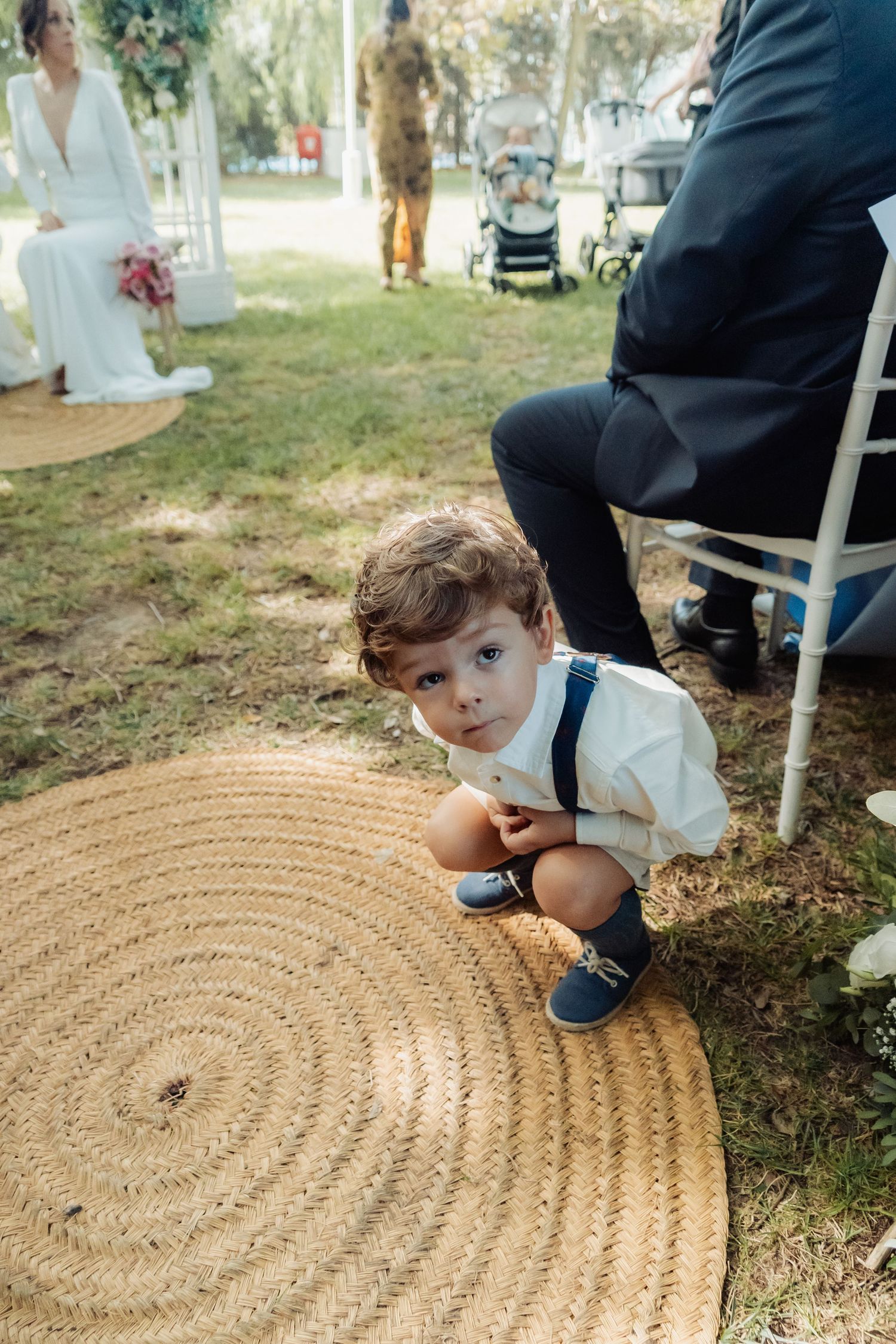 An adorable young boy in white shirt and suspenders crouches on a woven rug during an outdoor wedding ceremony.