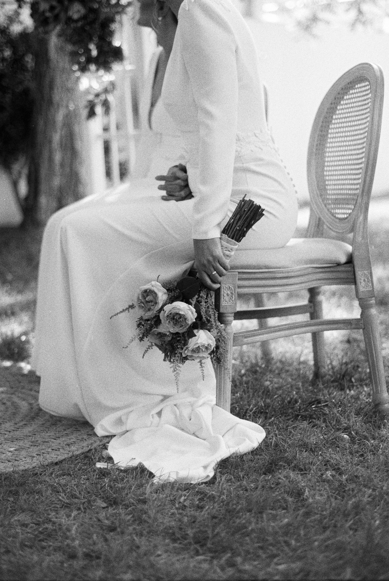 Black and white photo of seated bride in white gown holding bouquet of roses on chair.