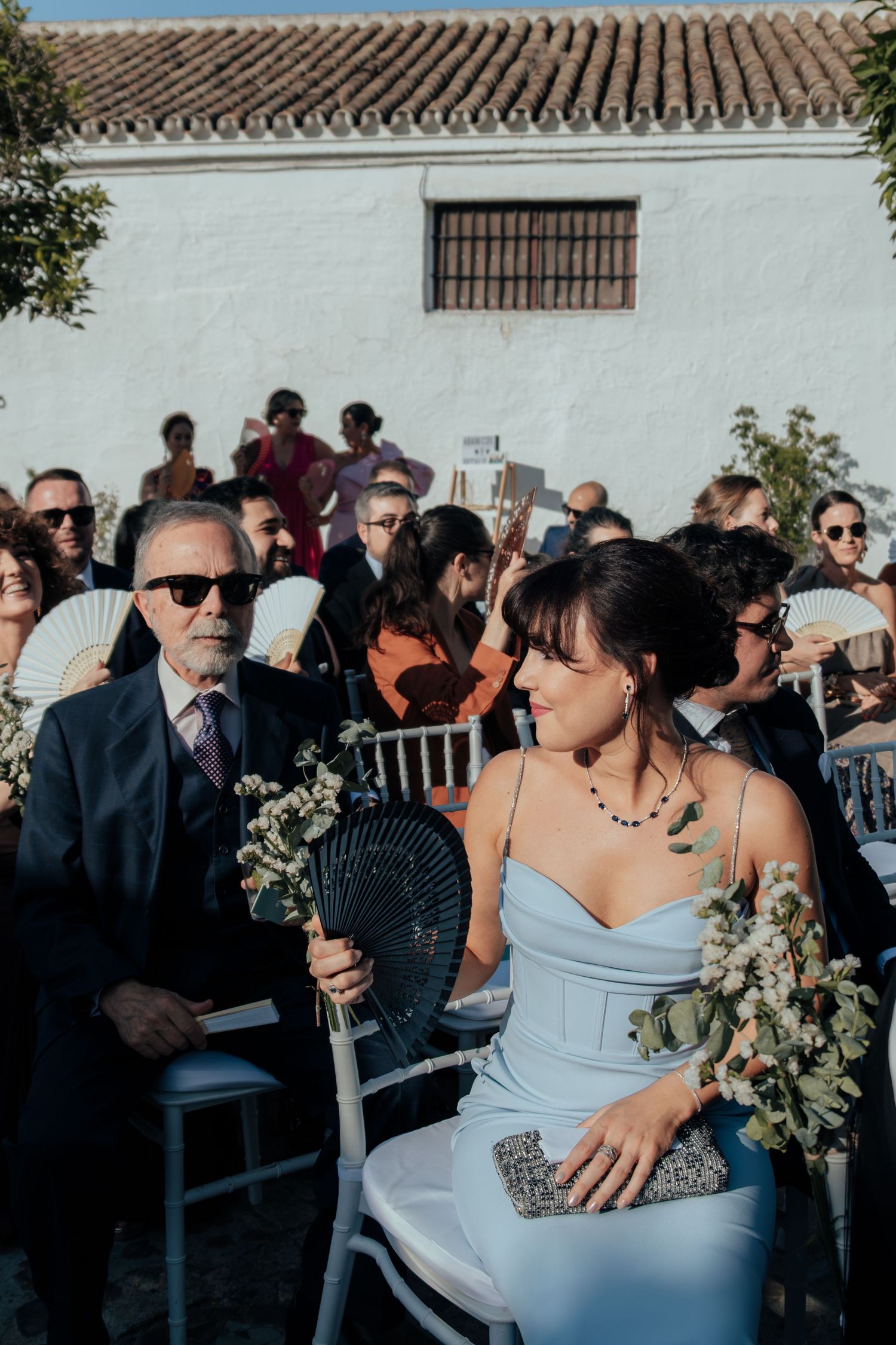 Elegantly dressed wedding guests seated in white chairs at an outdoor ceremony holding decorative fans.