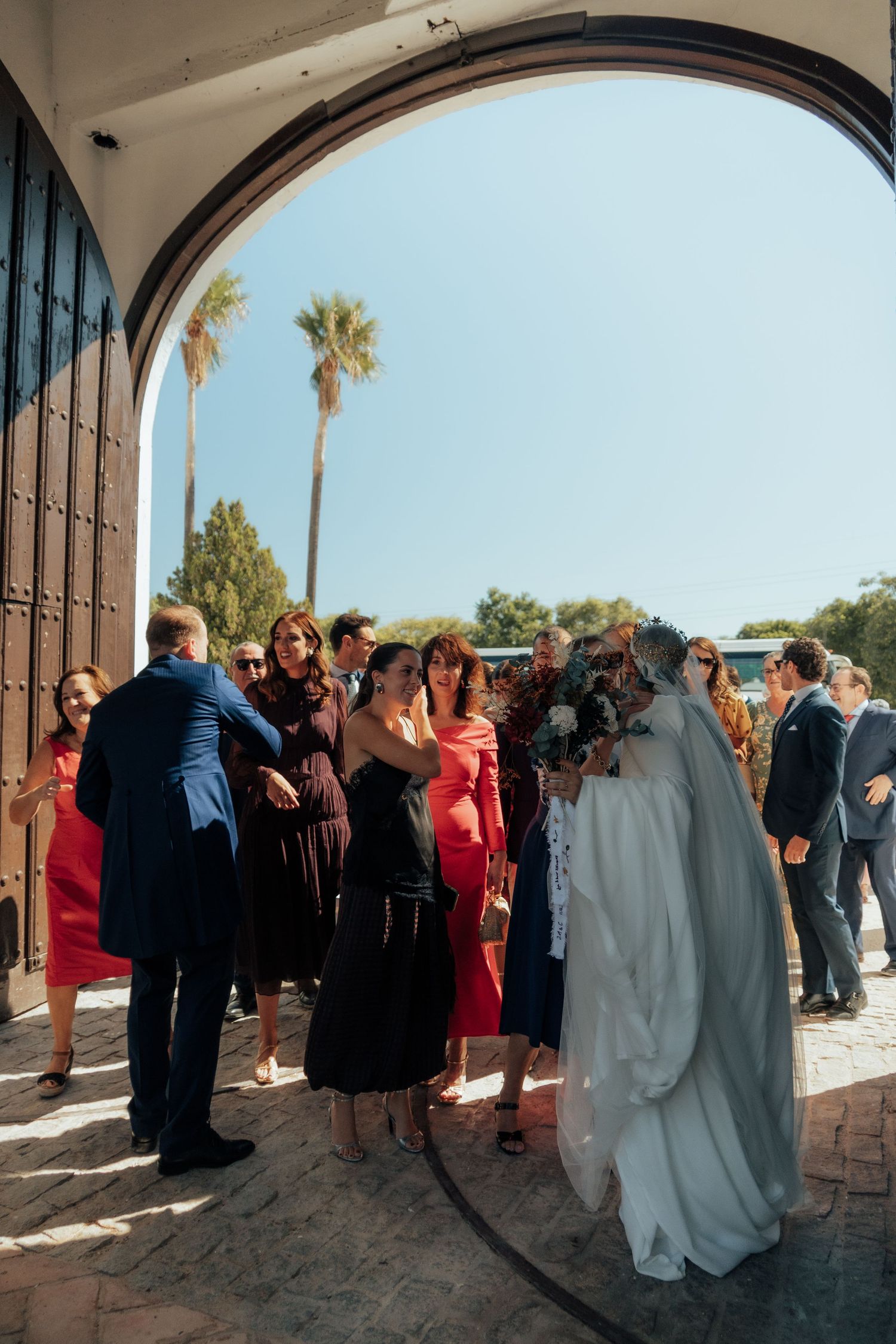 Wedding guests greet bride and groom under arched entrance of a sunny Spanish hacienda venue.