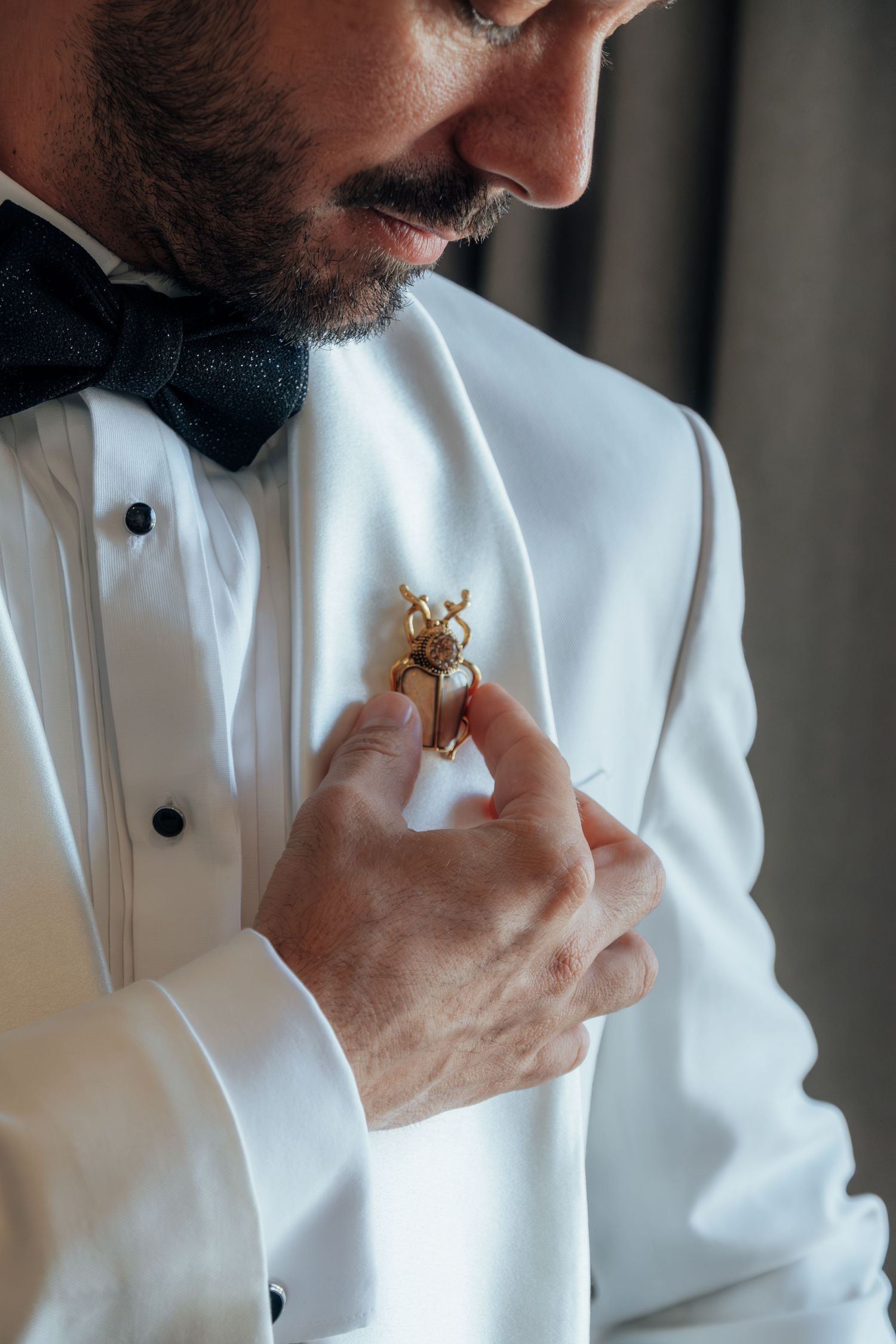 Close-up of a bearded groom in white tuxedo carefully pinning a gold beetle boutonniere.