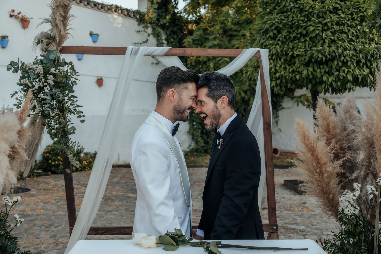 Two grooms share intimate forehead touch moment under decorated wedding arch with pampas grass.