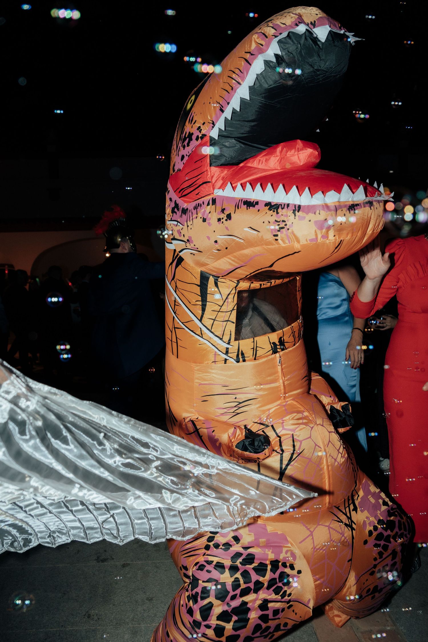 Person wearing an inflatable T-Rex dinosaur costume at a lively nighttime outdoor wedding celebration.