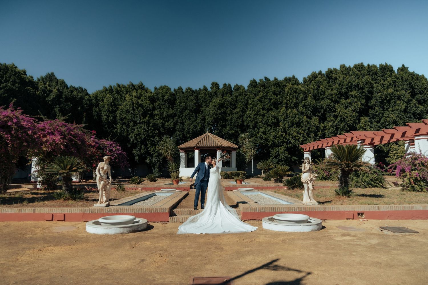 Bride and groom posing in a grand formal garden with statues and a classical pavilion backdrop.