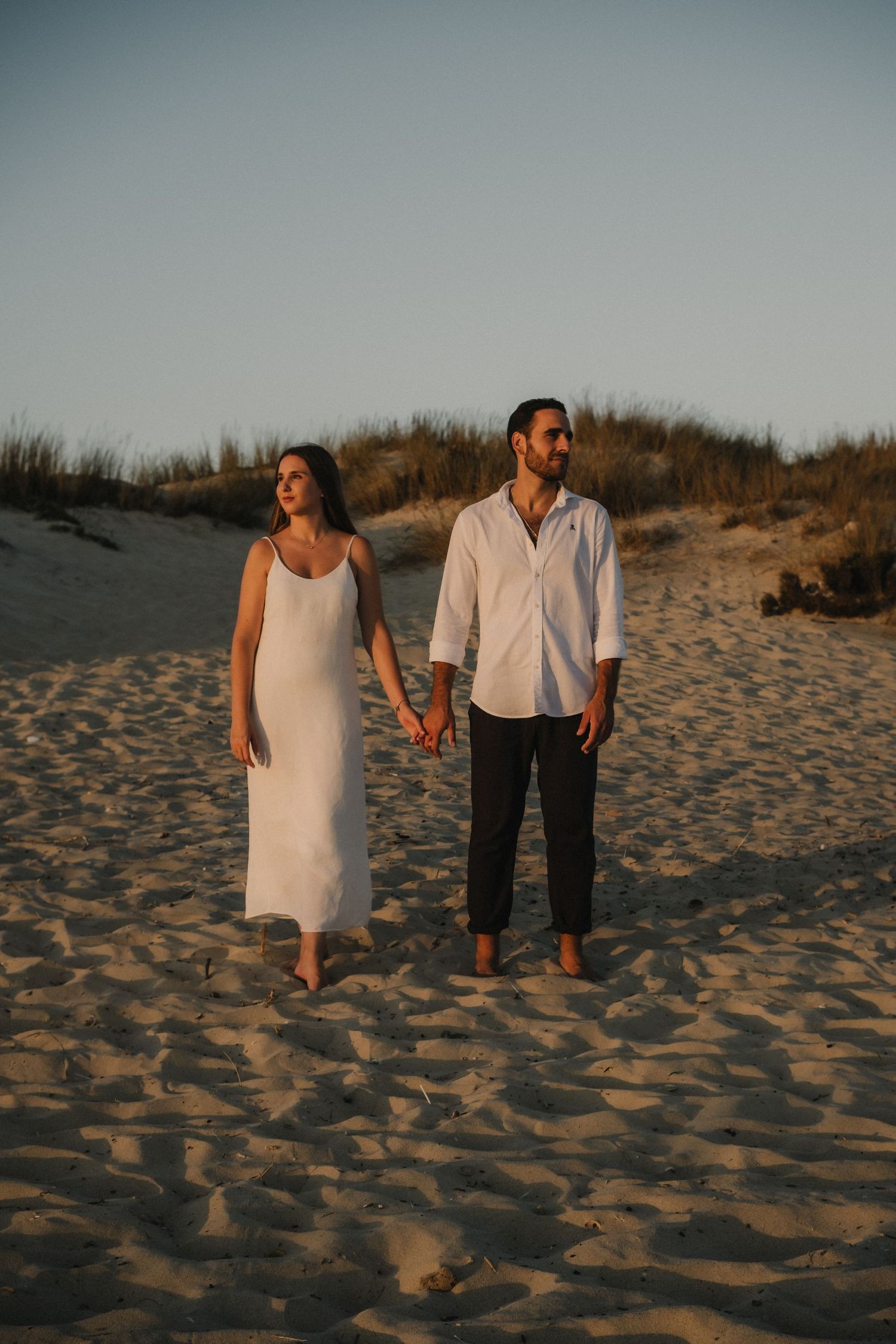 Couple holding hands walking barefoot on sandy beach at golden hour, woman in white slip dress, man in white shirt.