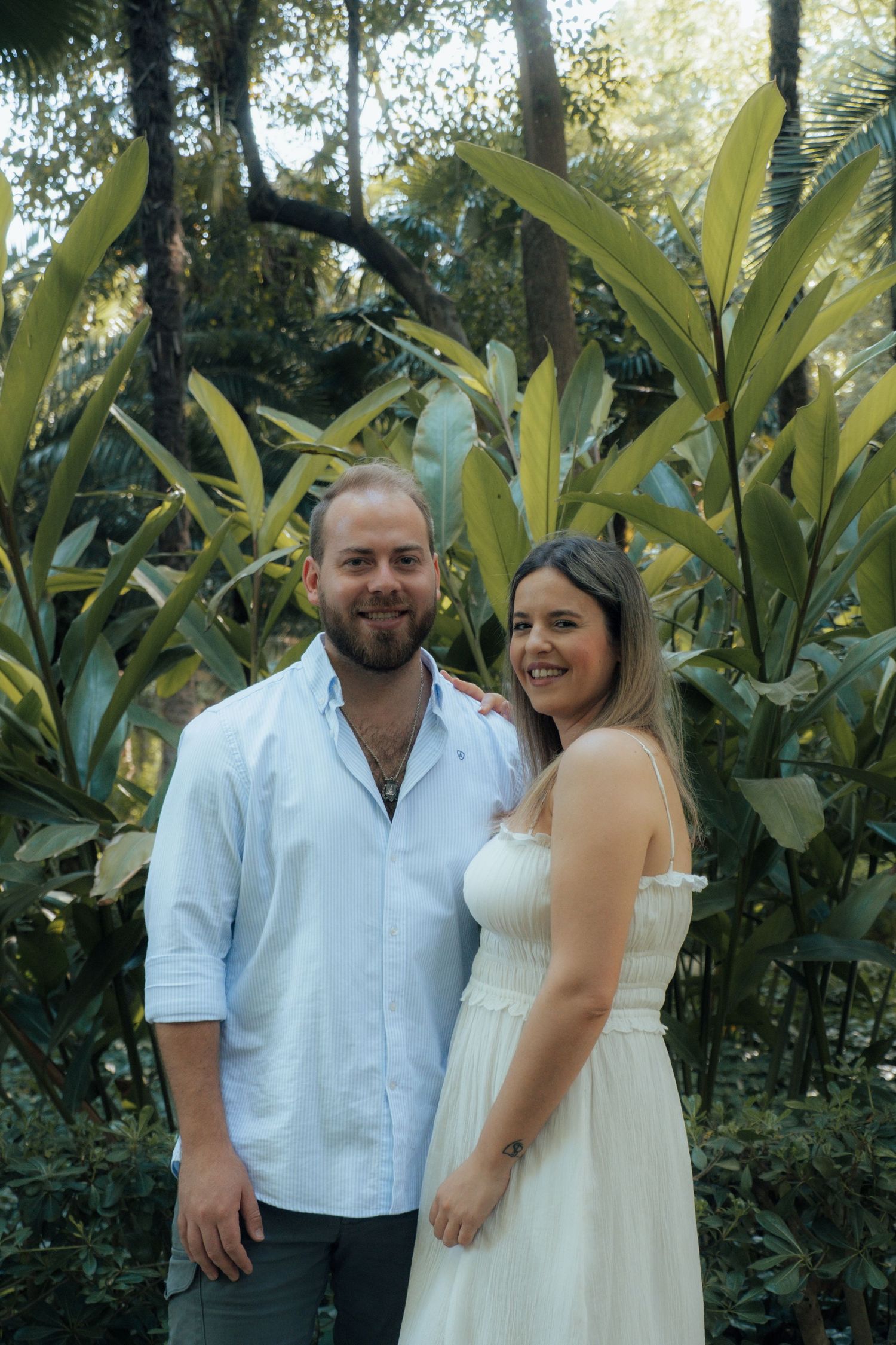 Smiling couple posing together before tropical foliage, man in blue shirt and woman in white sundress.