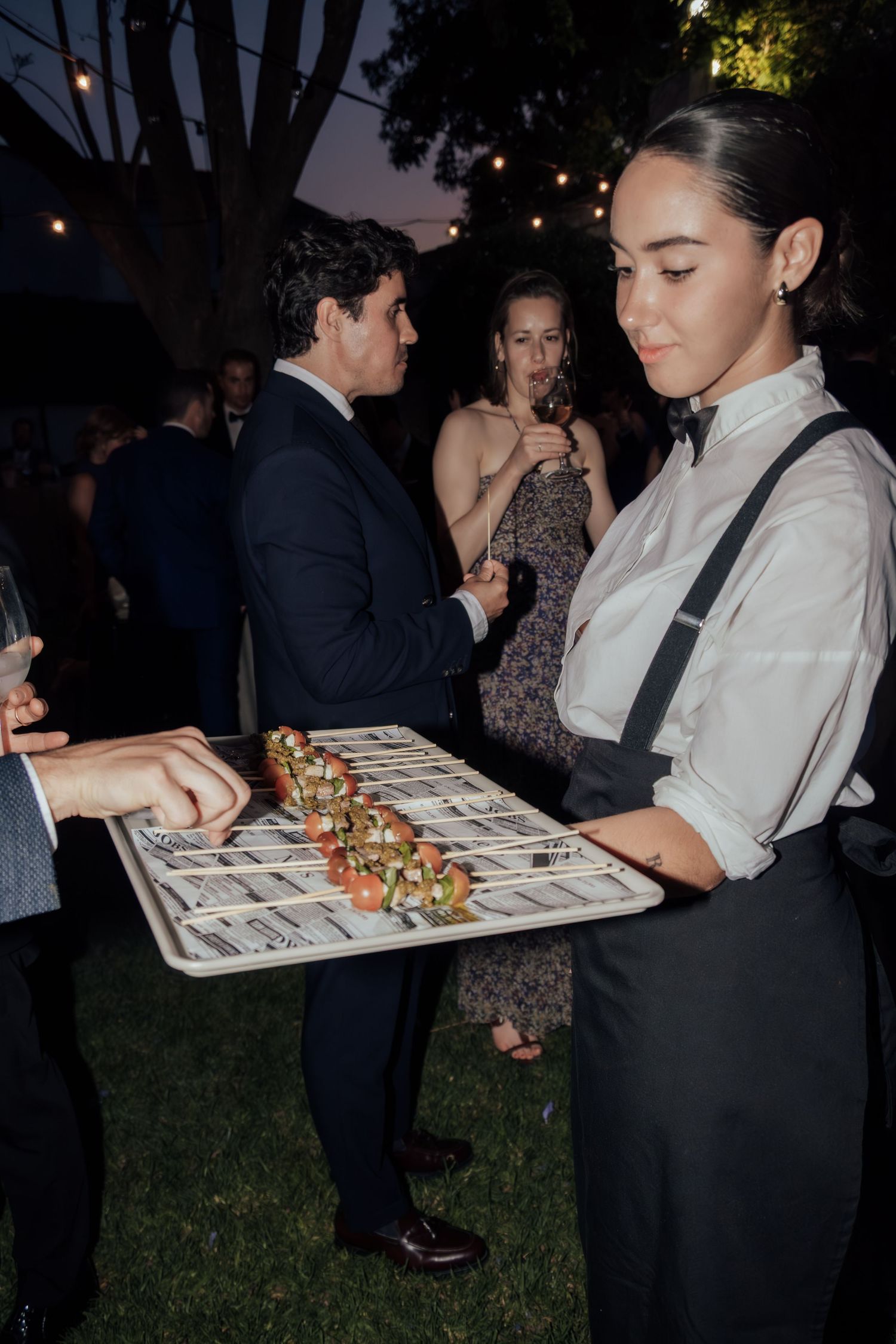 Elegant waitress in white shirt and suspenders serving skewer appetizers on tray at outdoor event.