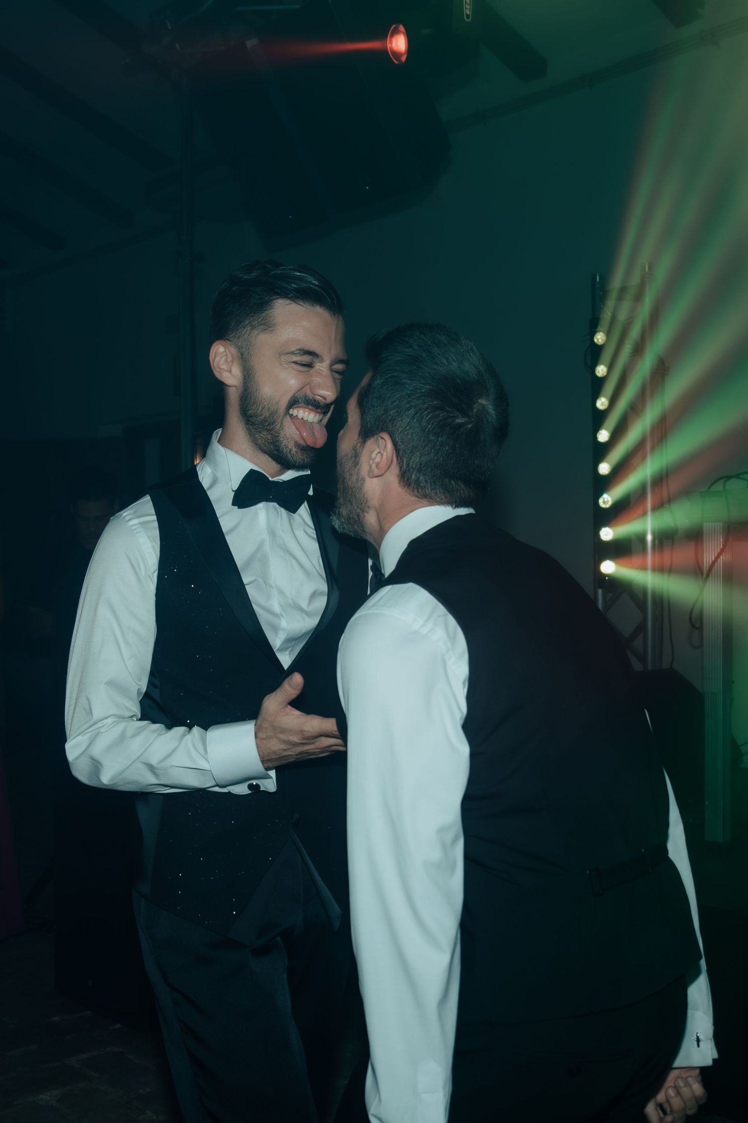Two grooms in black vests laughing and dancing together under colorful laser lights at wedding reception.