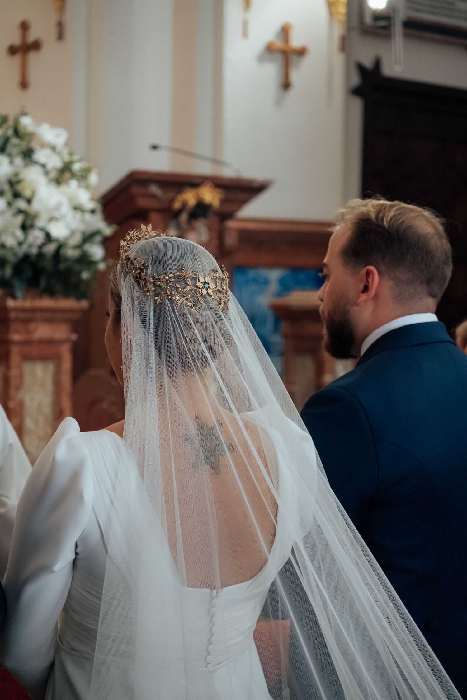Bride and groom stand at church altar, bride wearing white gown with gold crown veil during wedding ceremony.