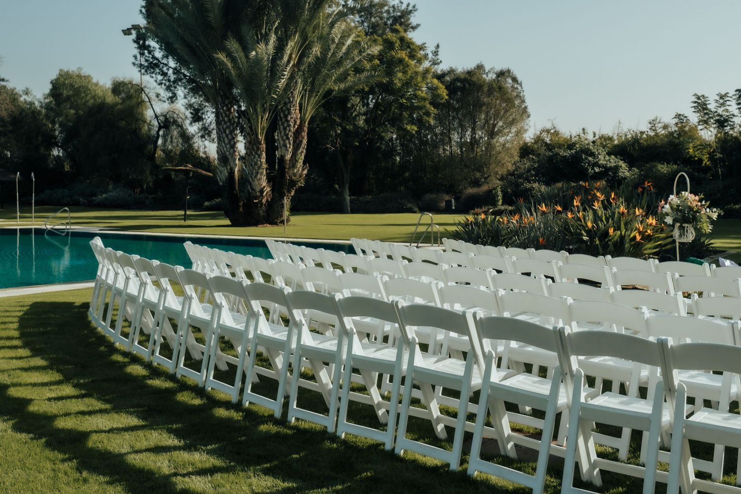 Rows of white folding chairs arranged on green lawn for outdoor wedding ceremony beside a pool.