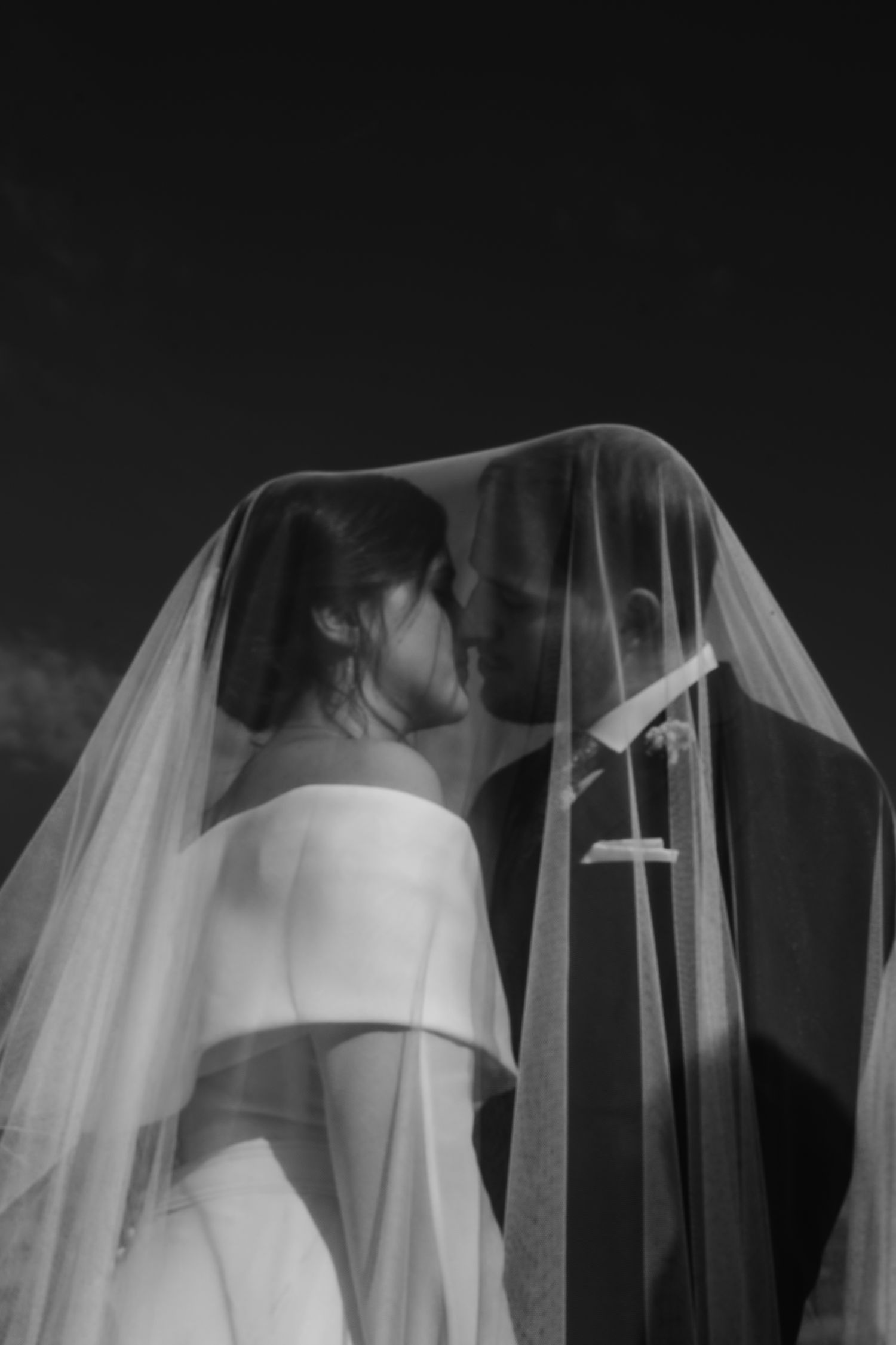 Black and white romantic photo of bride and groom kissing under a flowing wedding veil.