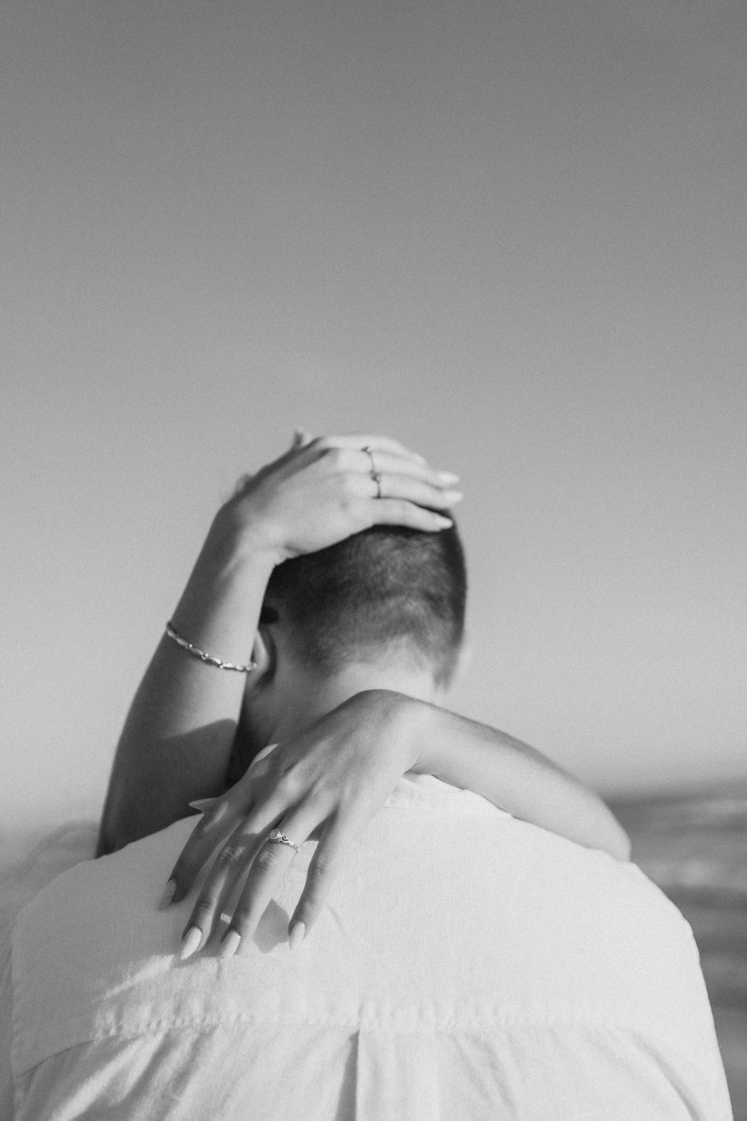 Black and white photo of a couple embracing, woman's hands with rings and bracelet resting on man's head and shoulder.