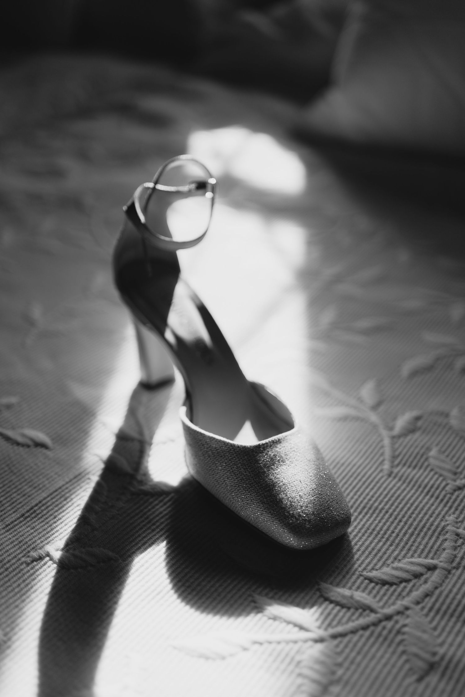 Black and white photo of a glittery high heel shoe with ankle strap casting dramatic shadow in sunlight.