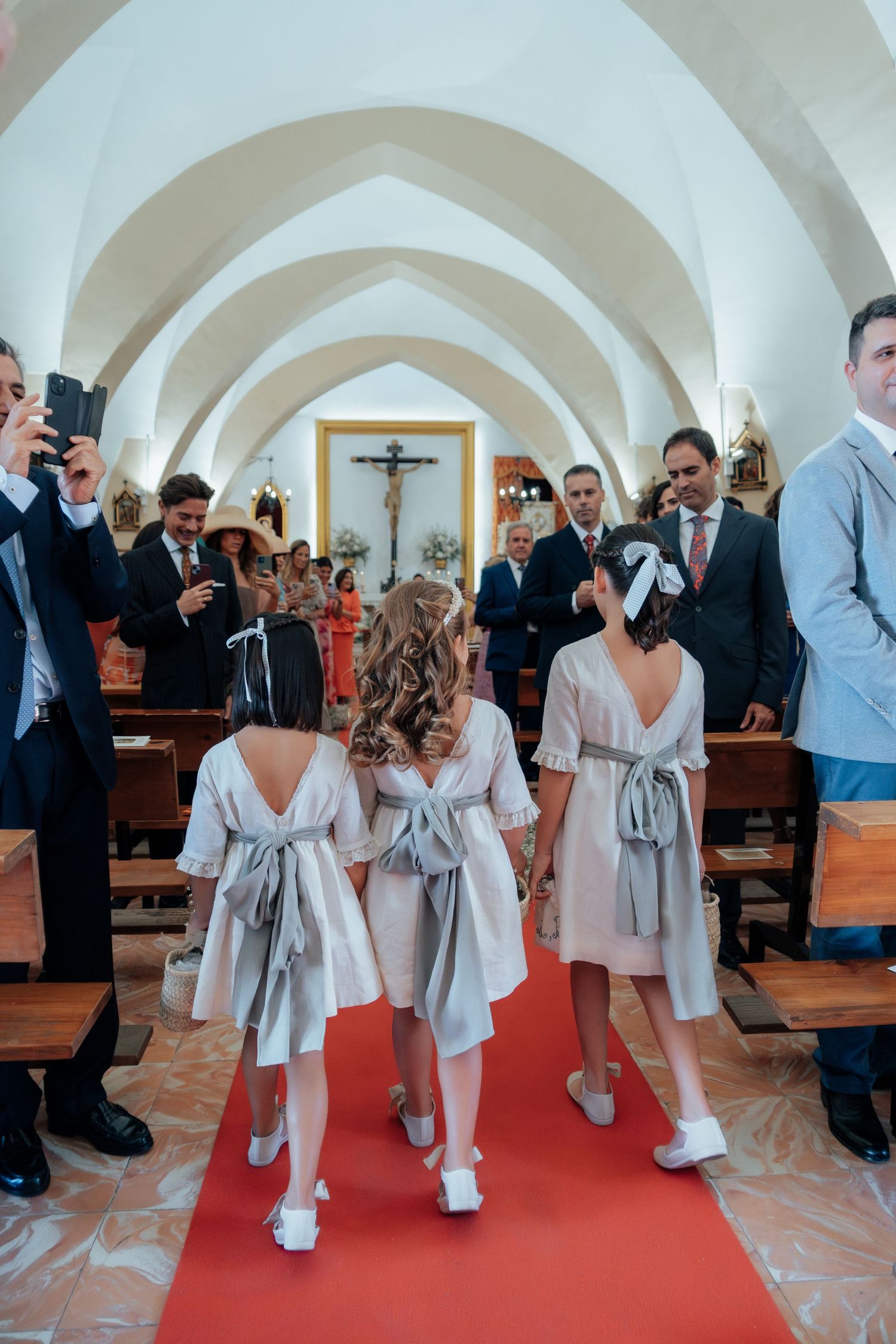 Three flower girls in matching grey dresses walk down red carpet church aisle at wedding.