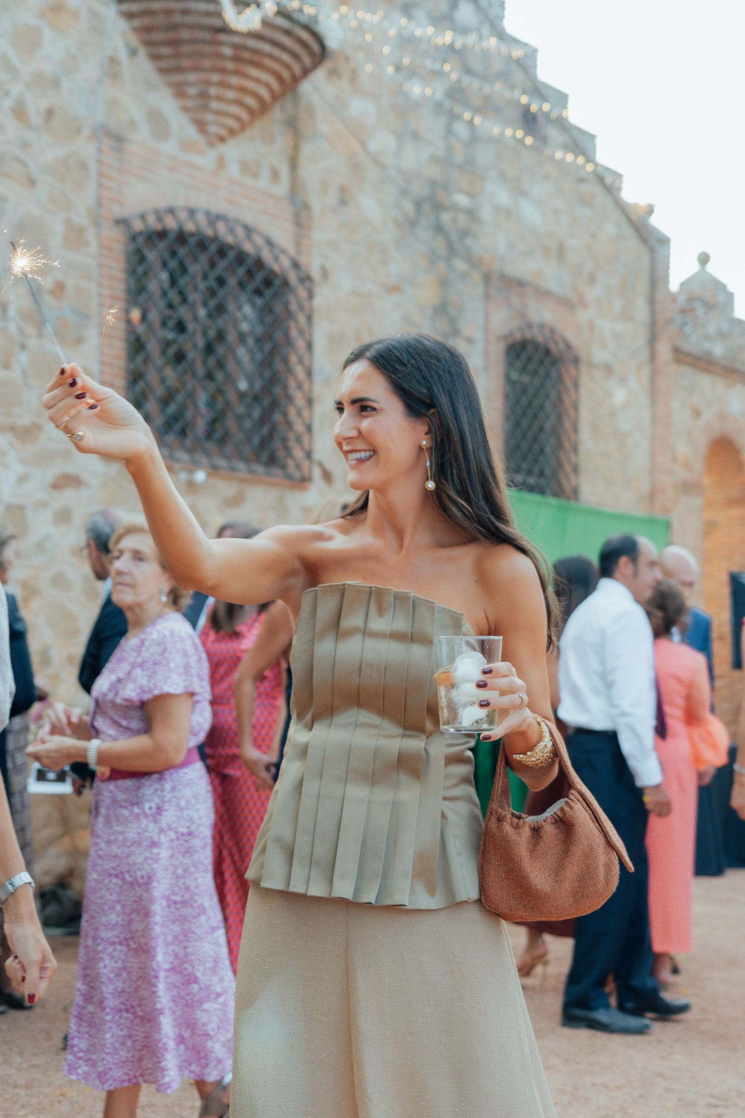 Elegant woman in strapless olive dress holding sparkler and drink at outdoor wedding celebration.
