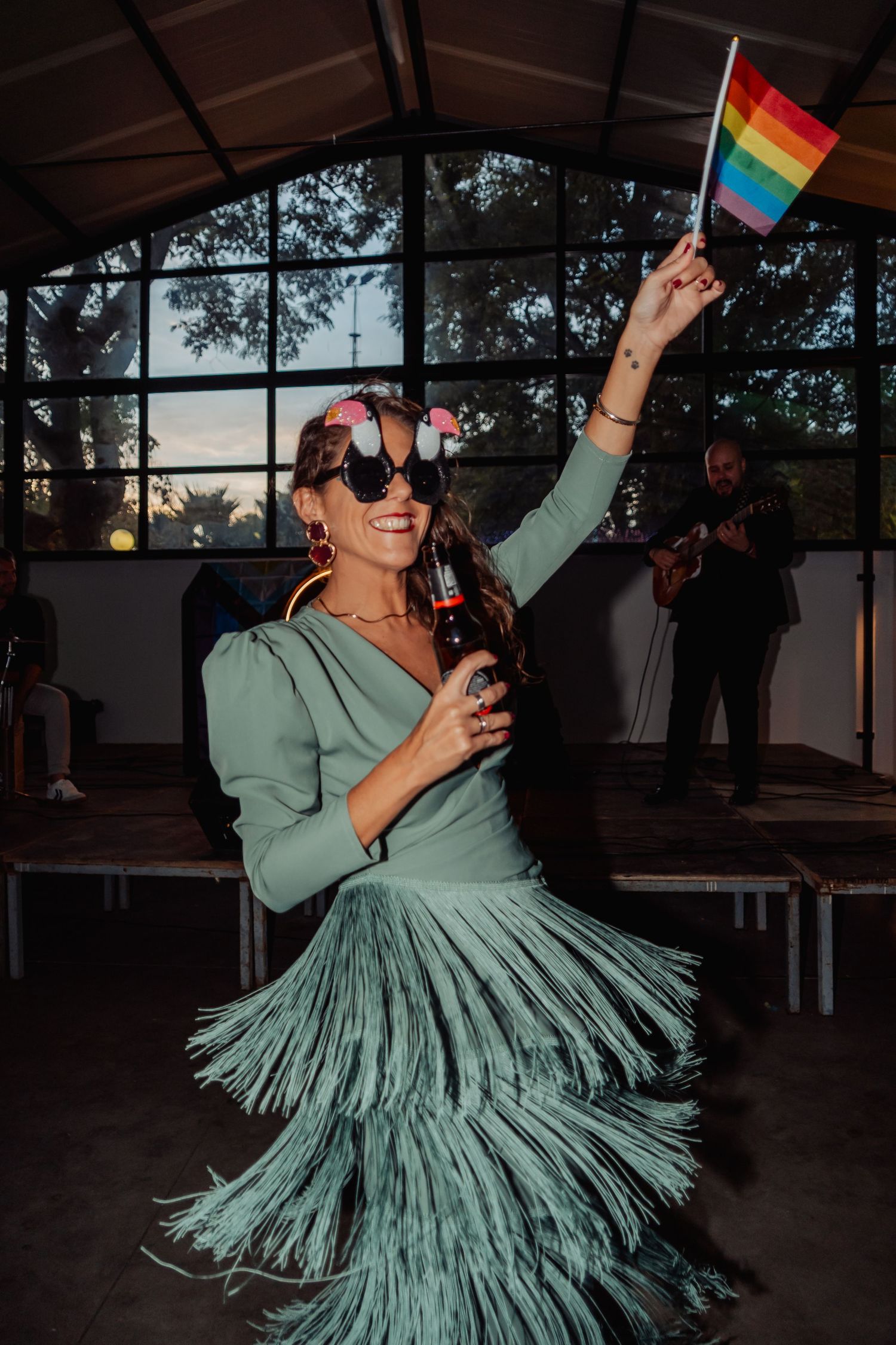 Woman in green fringe dress waving rainbow pride flag while dancing at an outdoor celebration.