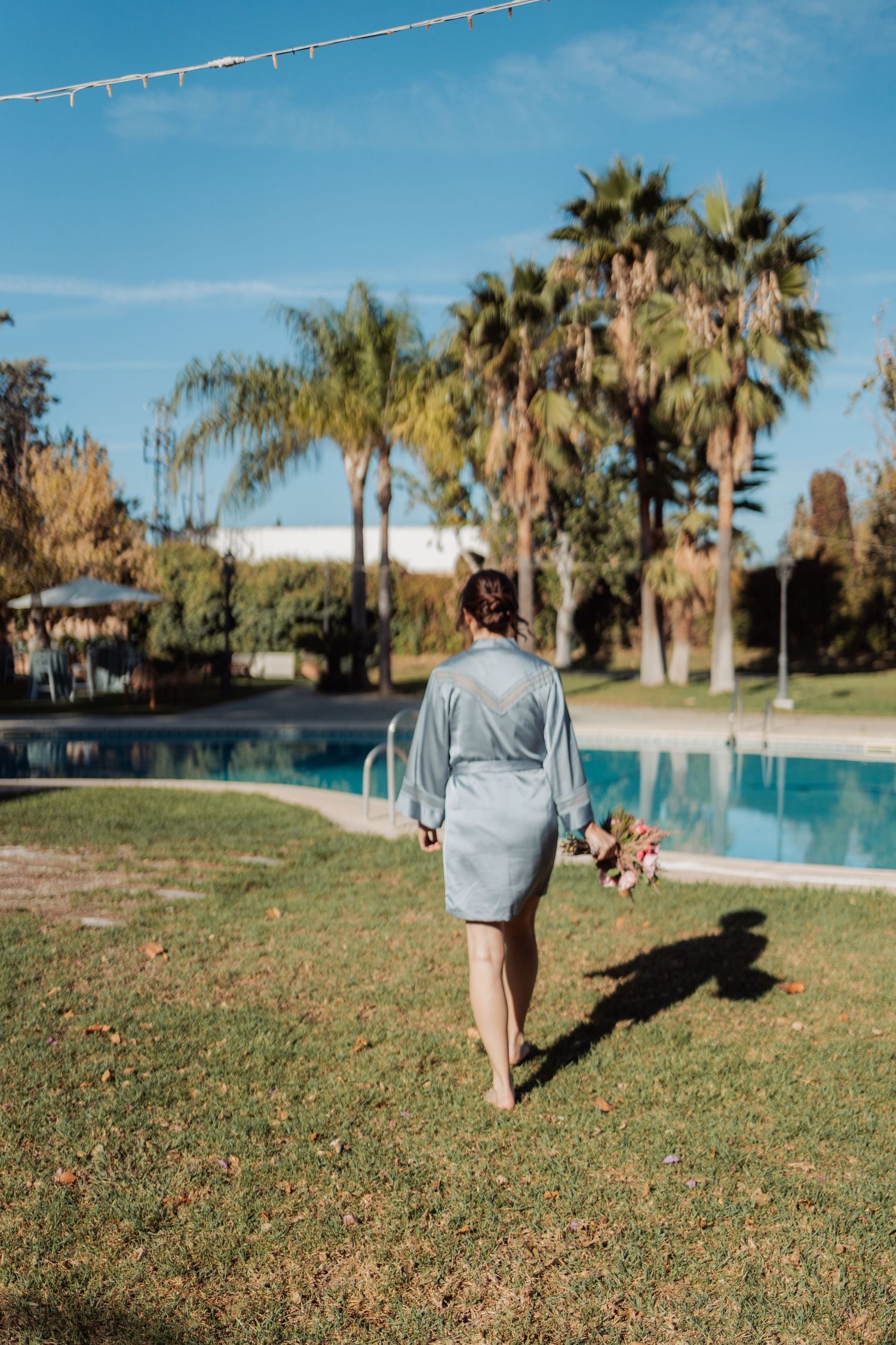 Barefoot woman in blue robe walks across green lawn holding bouquet near pool with palm trees.