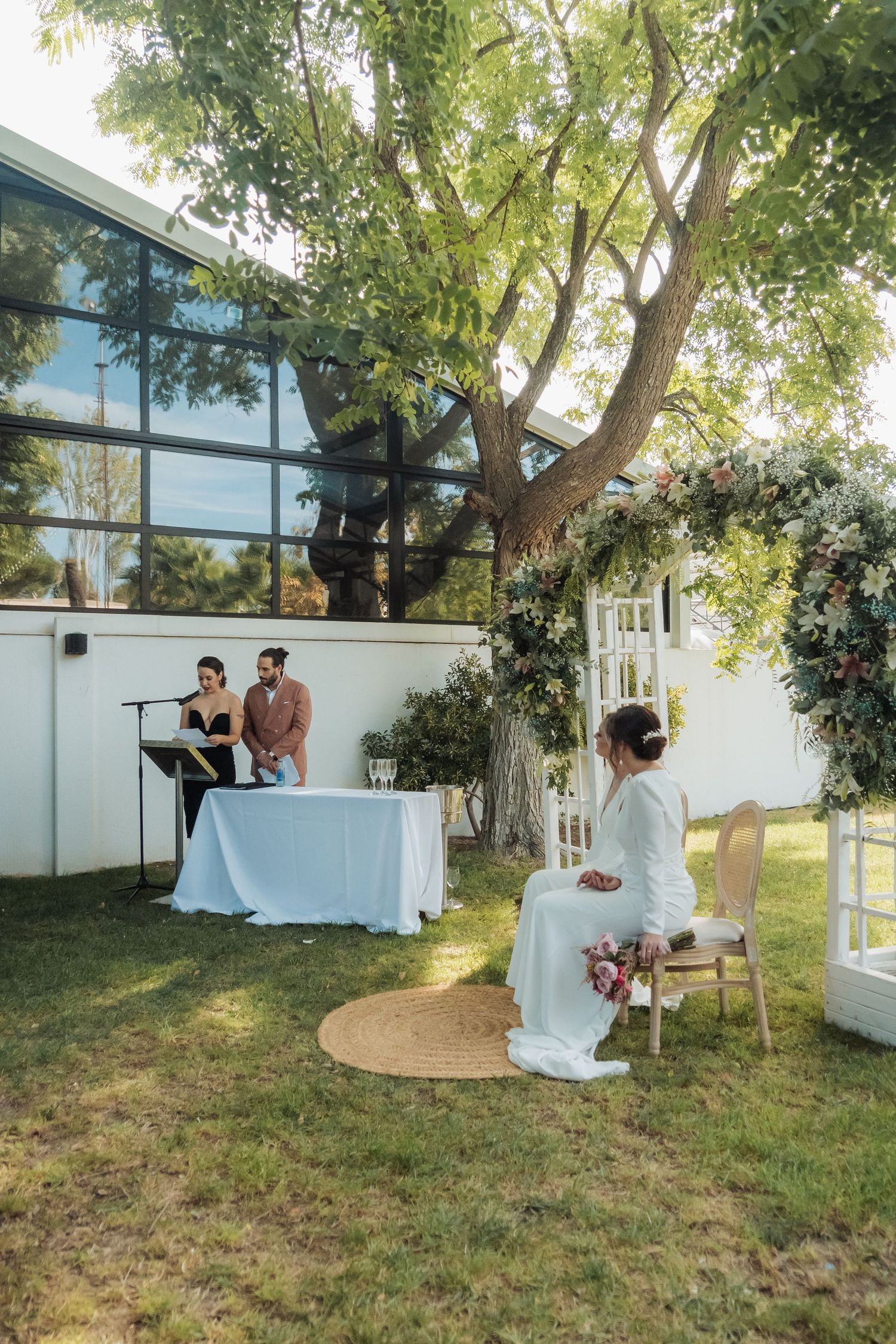 Outdoor wedding ceremony with bride seated before officiant at a white-draped table in a garden.