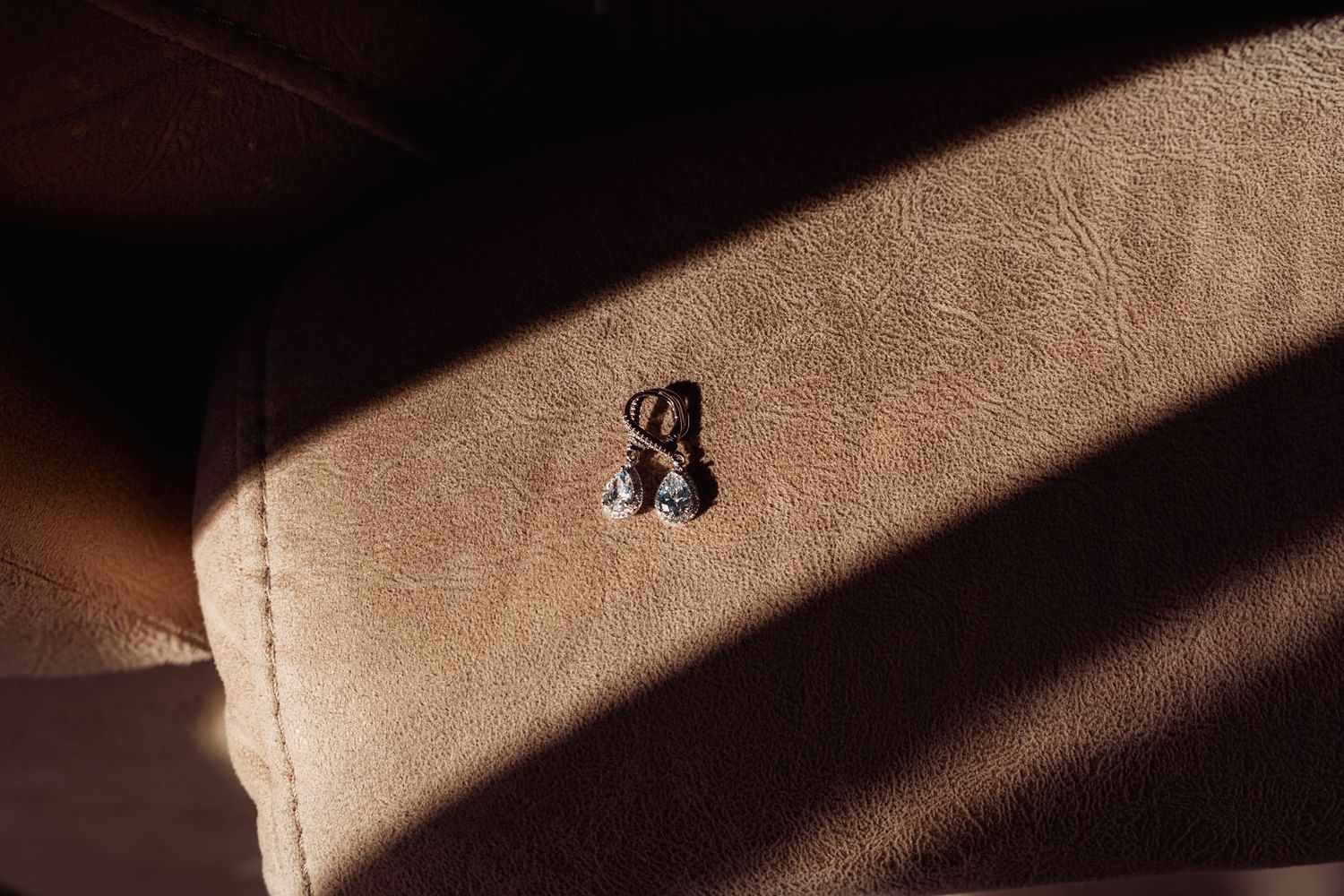 Diamond drop earrings resting on tan leather surface with dramatic sunlight shadow.