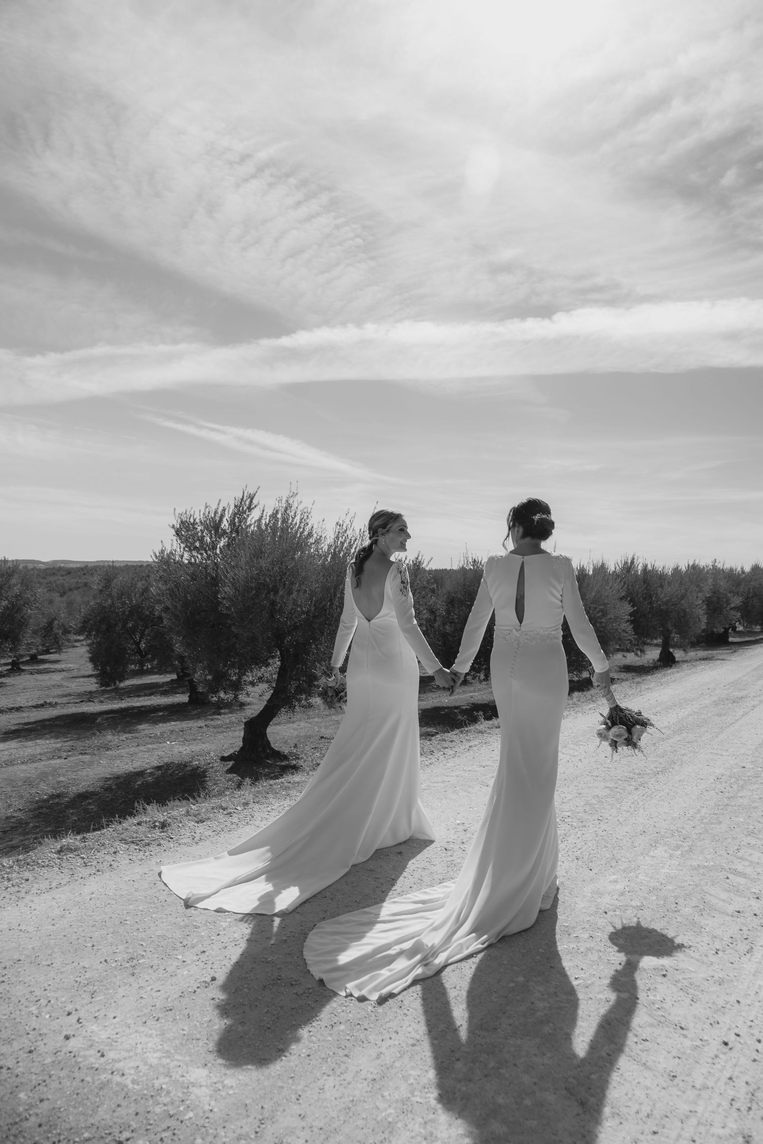 Black and white photo of two brides holding hands on a rural dirt road through olive trees.