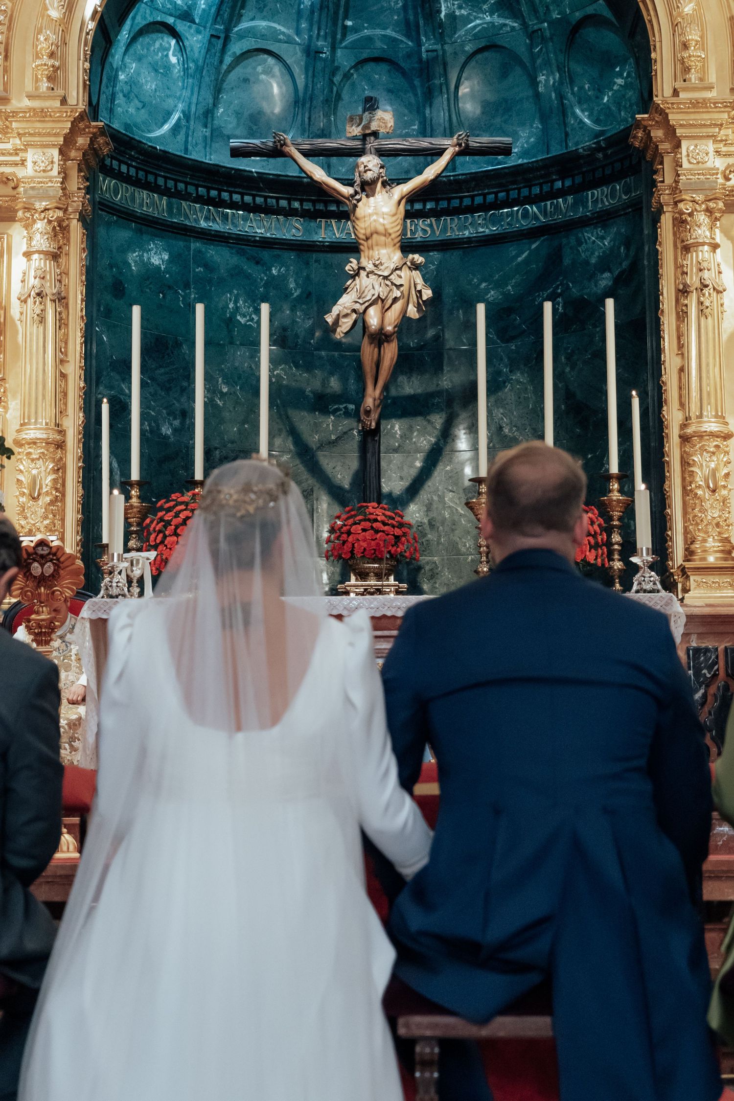 Bride and groom stand before ornate golden altar with crucifix during Catholic church wedding ceremony.