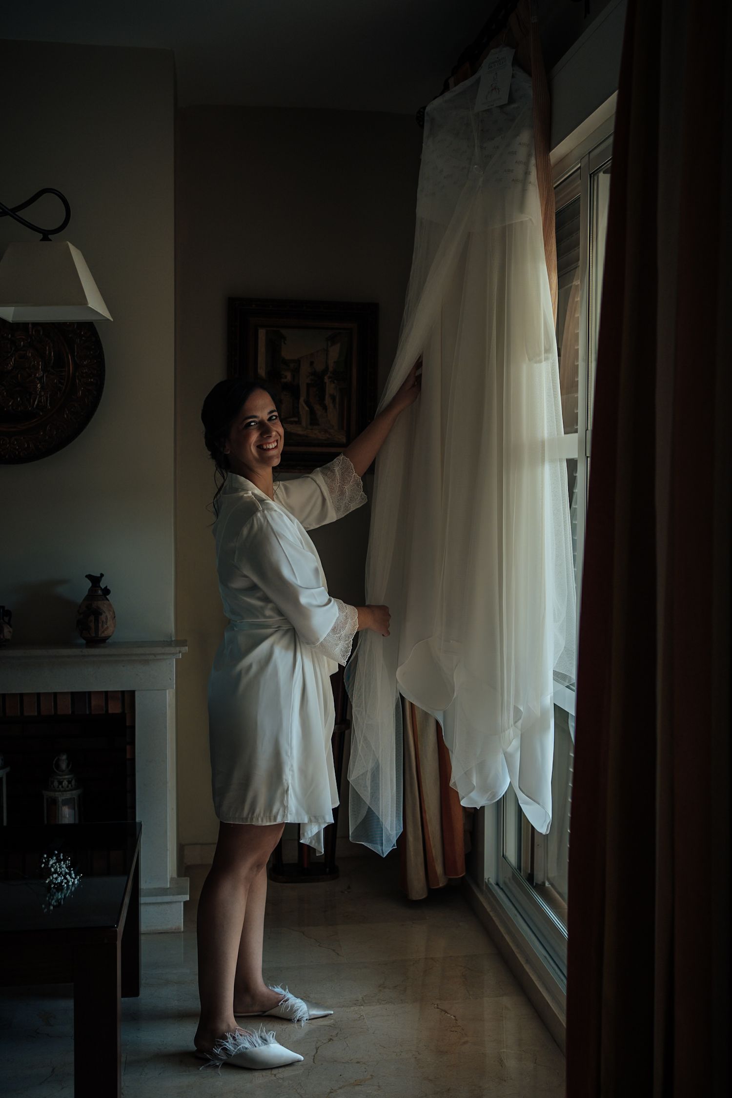 Smiling bride in white robe reaching for her wedding gown hanging by a sunlit window indoors.