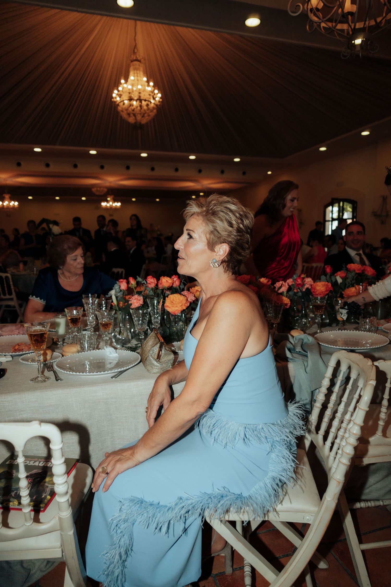 Woman in light blue feathered dress seated at wedding reception table with floral centerpieces.