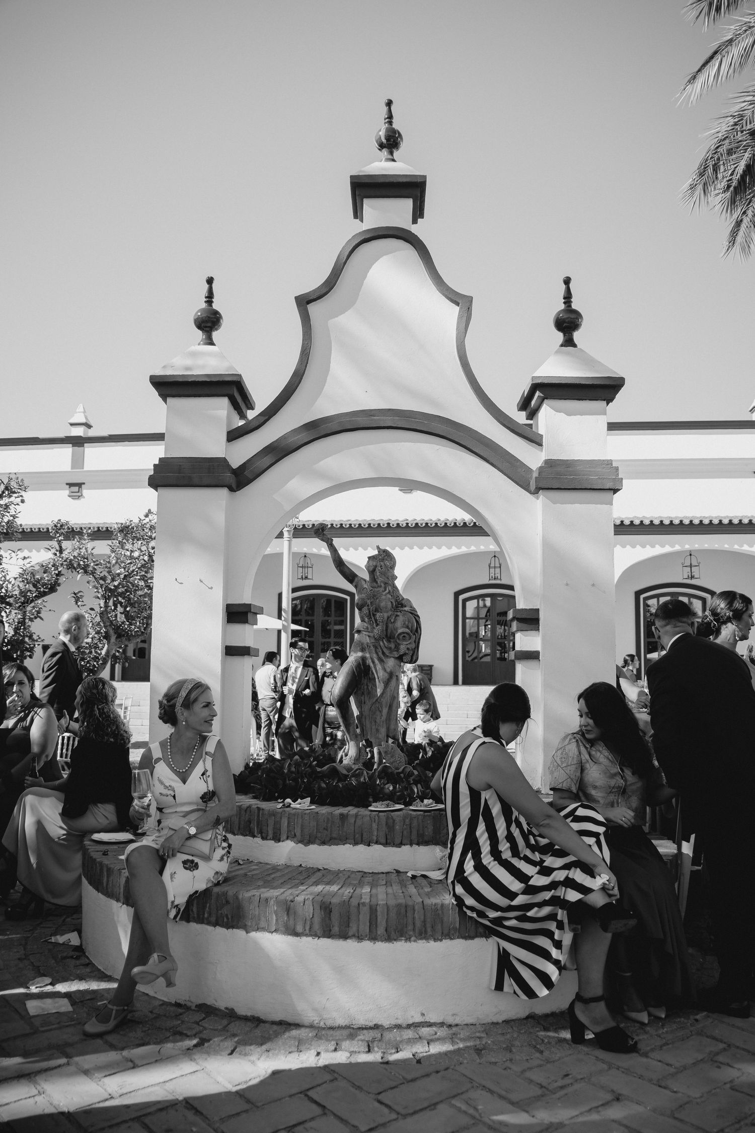 Black and white photo of wedding guests gathered around an ornate fountain at a Spanish venue.