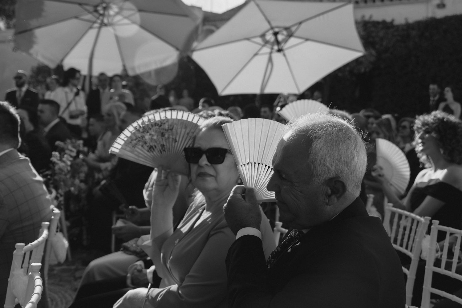 Wedding guests use decorative hand fans to stay cool during an outdoor ceremony in black and white.