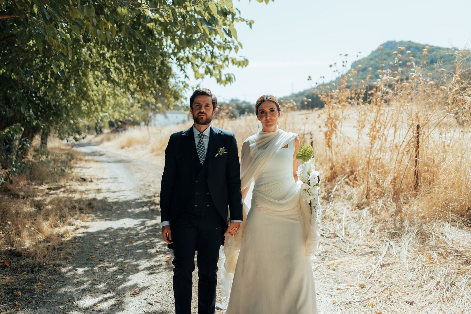 Bride and groom walking hand in hand on a rustic dirt path surrounded by golden dry fields.
