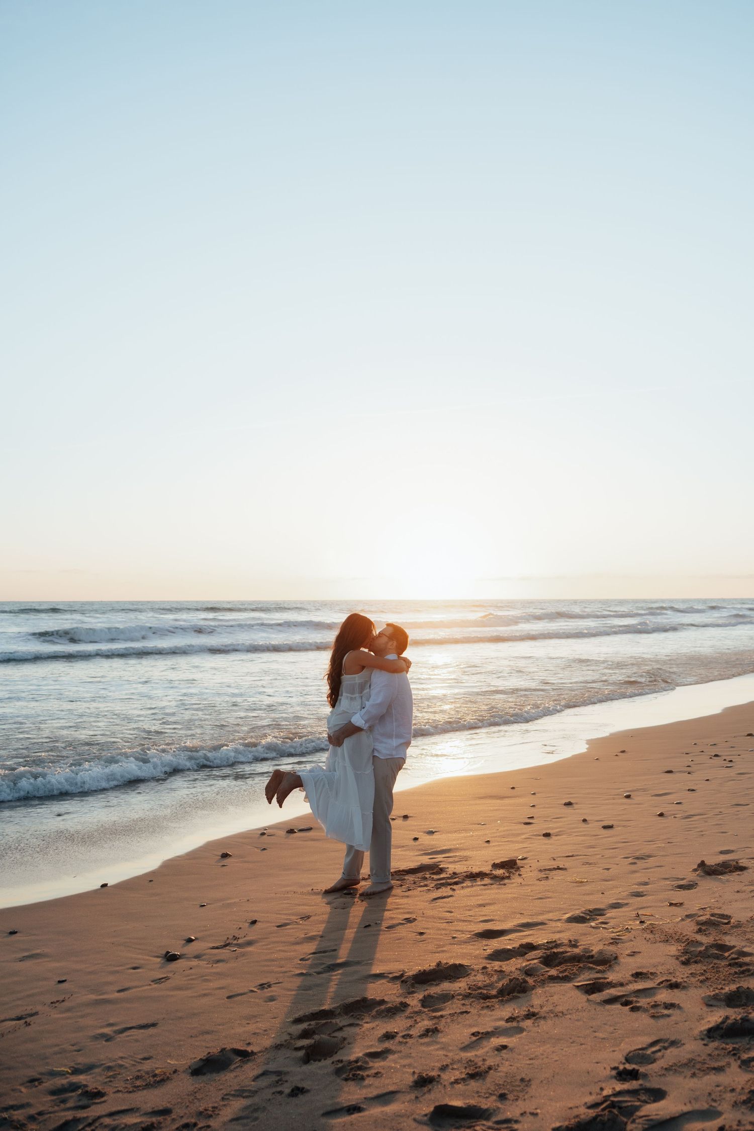 Couple embracing and kissing on a sandy beach at sunset with ocean waves in the background.