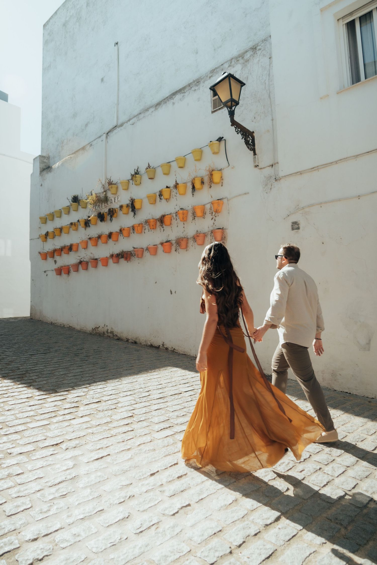 Couple walking hand-in-hand on cobblestone street past white wall with colorful flower pots in sunny European town.