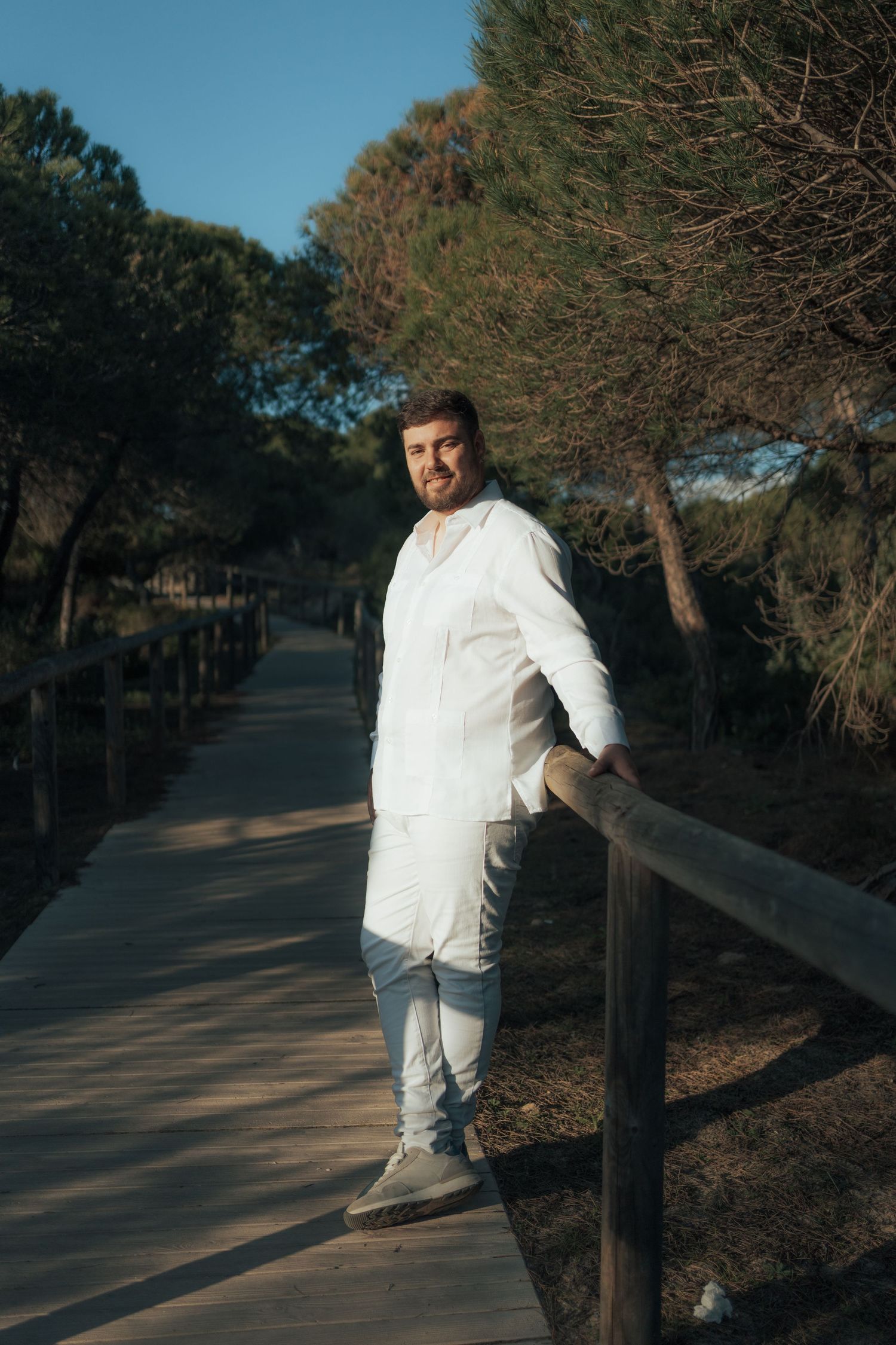 Man in white shirt and jeans leaning on wooden railing of a sunlit park boardwalk surrounded by pine trees.