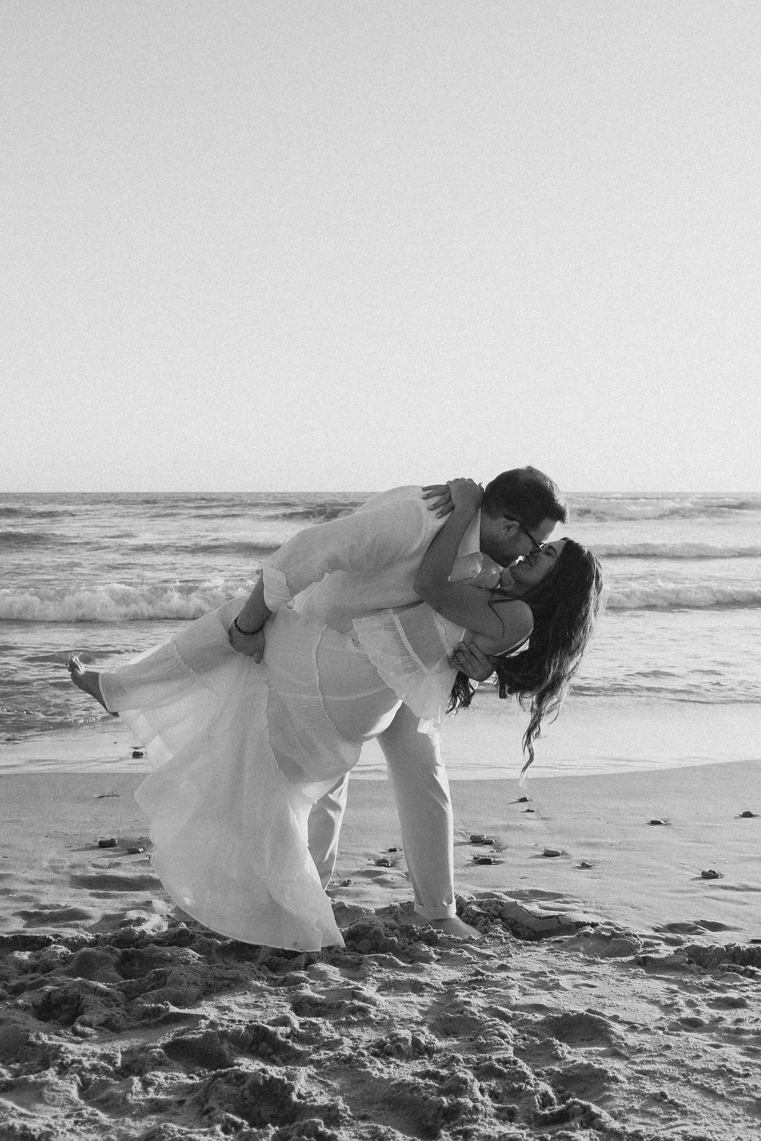 Black and white photo of a couple sharing a romantic dip kiss on a sandy beach with ocean waves behind them.