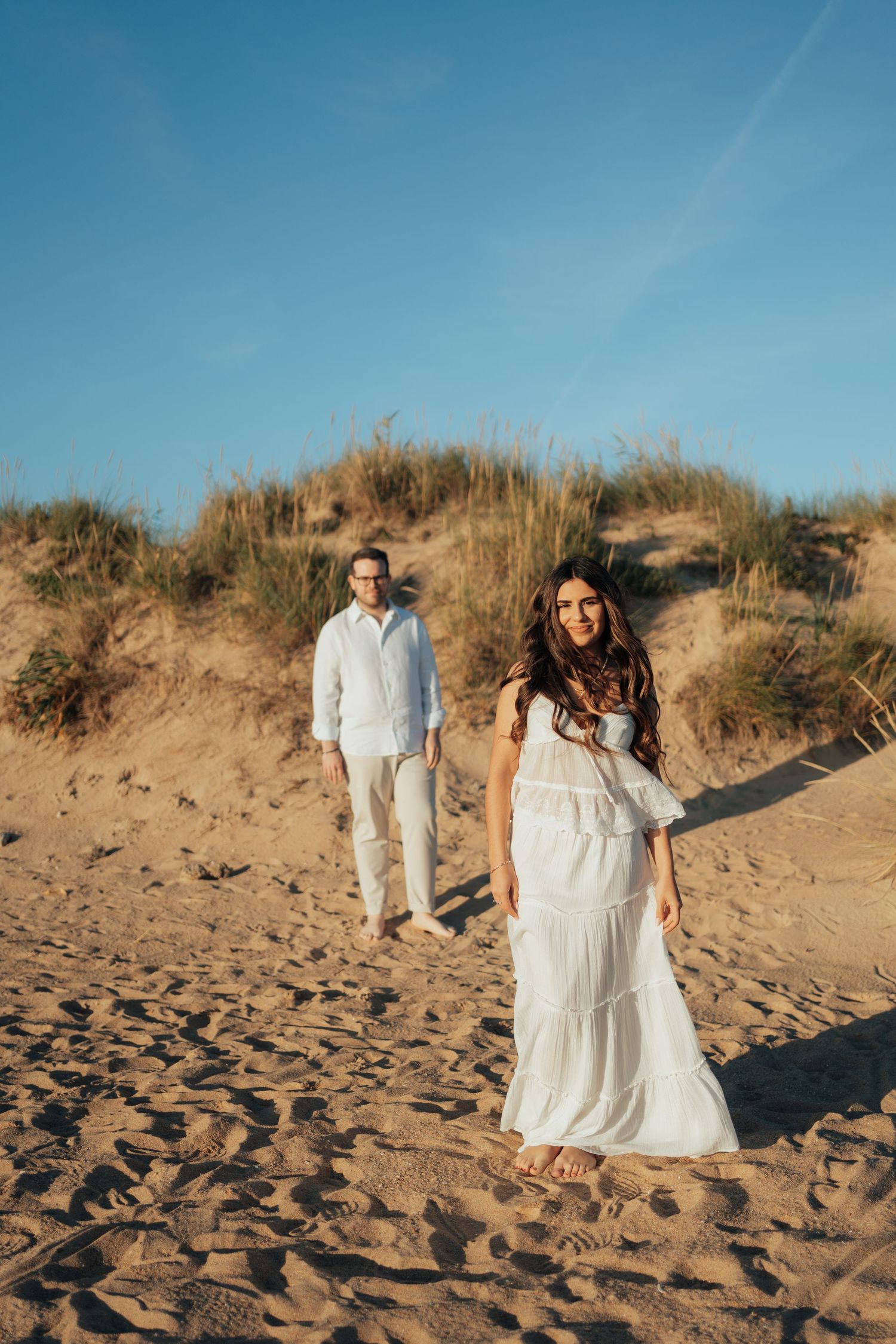 A couple walks on sandy beach dunes, woman in white dress foreground, man in white shirt behind, blue sky backdrop.
